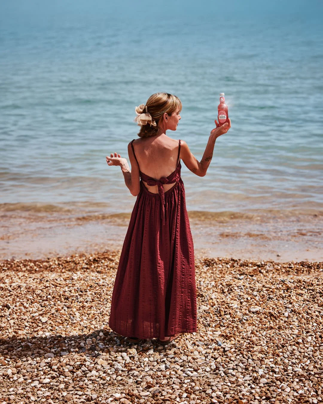 girl looking out to sea in stunning summer beach scene in red dress holding bottle of rhubarb lemonade