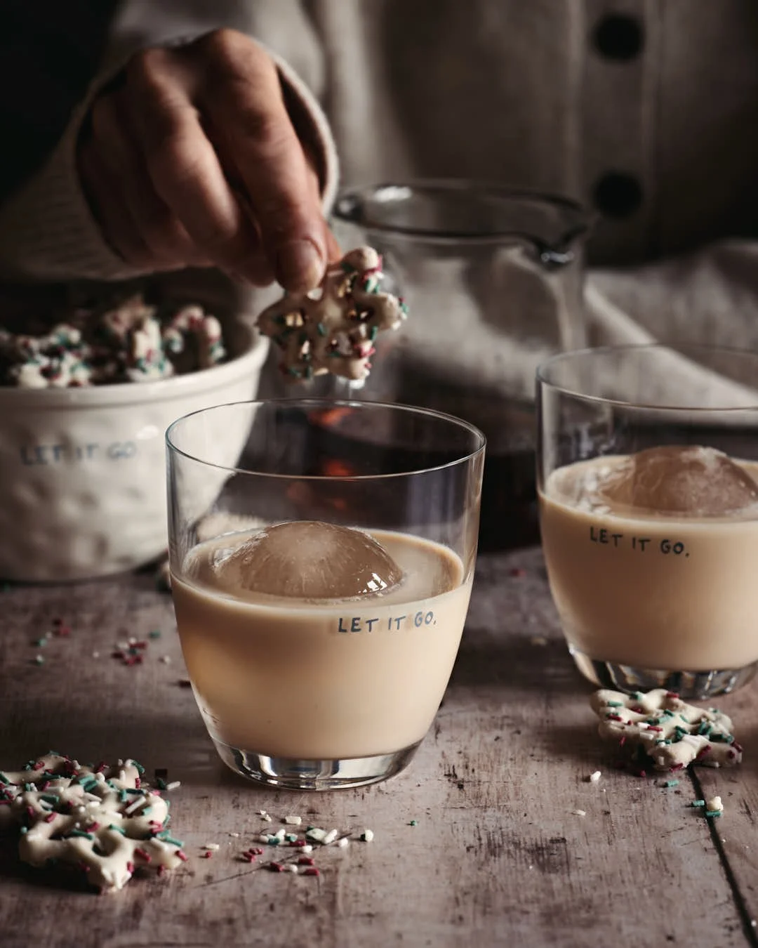 girl's hand leaning in to dunk white chocolate pretzel into glass of rum cream in festive winter scene on worn wood table