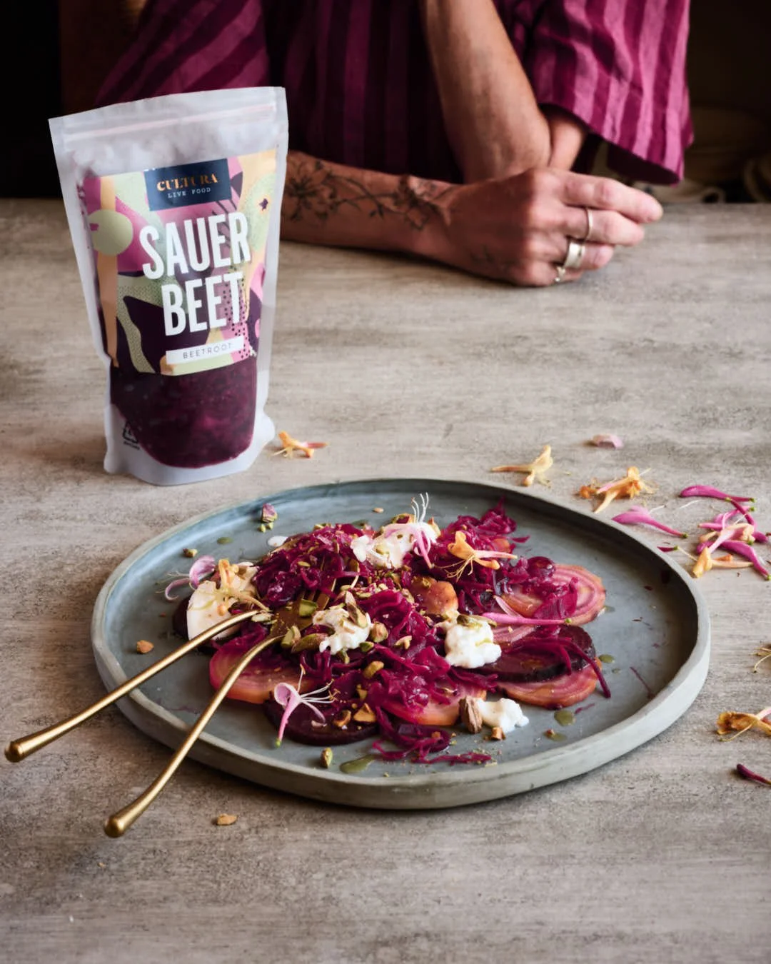 Angled image of beet salad with burrata and pistachio texture in bright scene with girl in deep pink striped dress sitting to back of bag of beet kraut