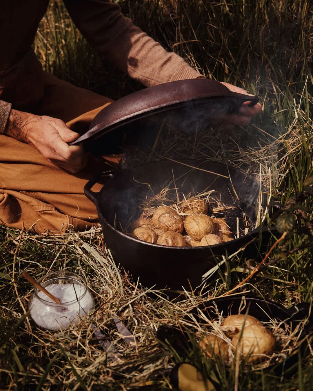 angled image of potatoes nestled in hay in cast iron pot with salt being sprinkled in outdoor setting