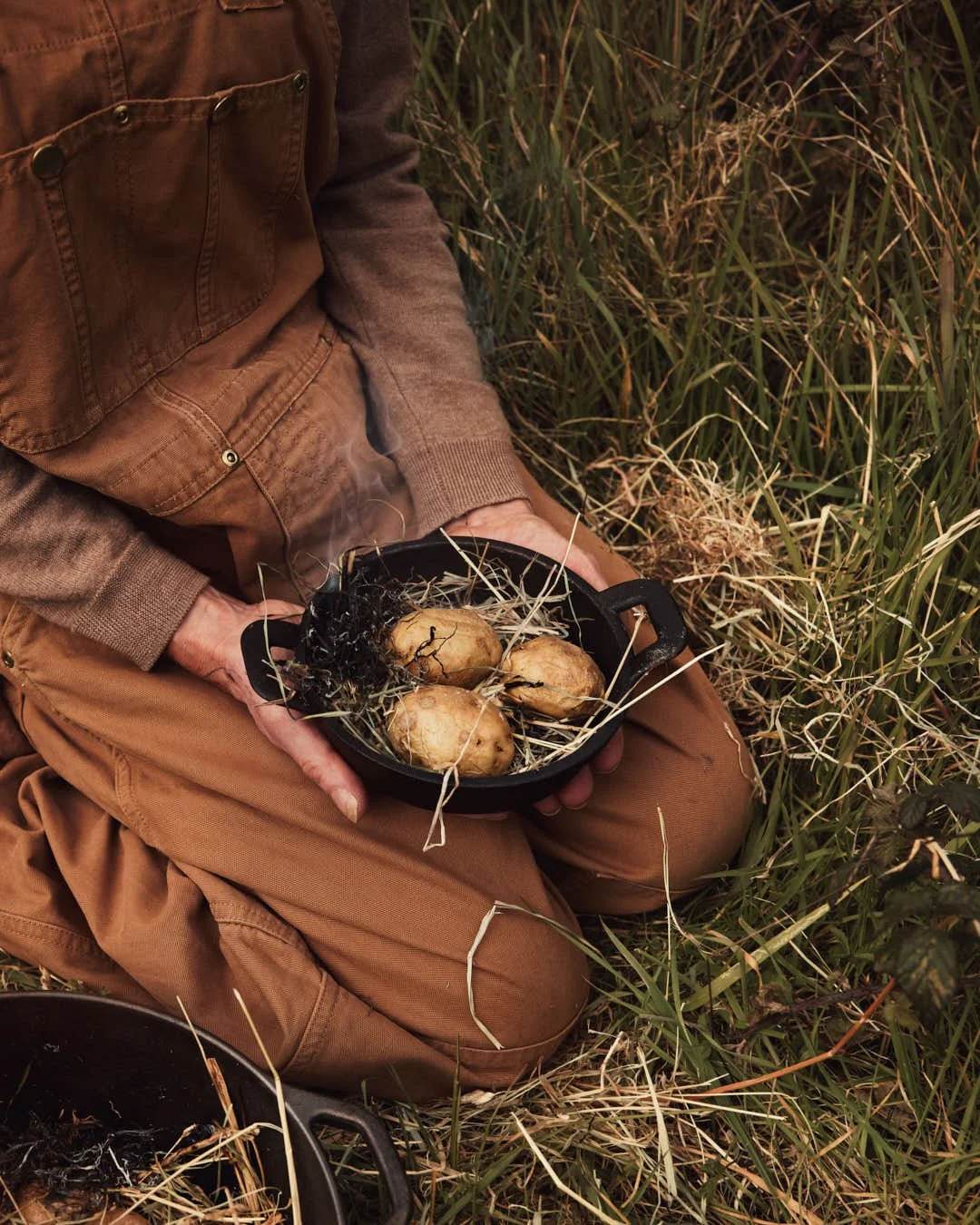 girl holding angled image of potatoes nestled in hay in cast iron pot with salt being sprinkled in outdoor setting