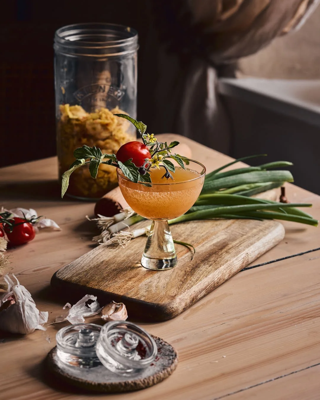 angled straight on image of glass jar of kimchi with chopping board and ferment presser in farmhouse scene with kimchi martini in focus