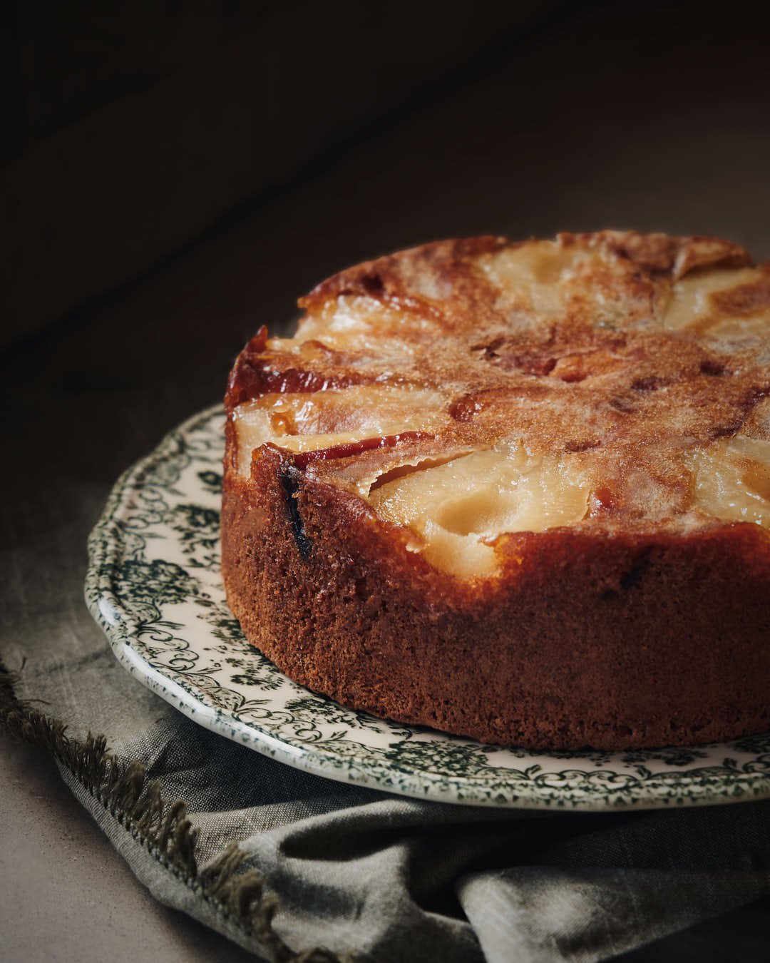 stunning upside down cake surrounded by pears on green patterned plate in moody lit scene