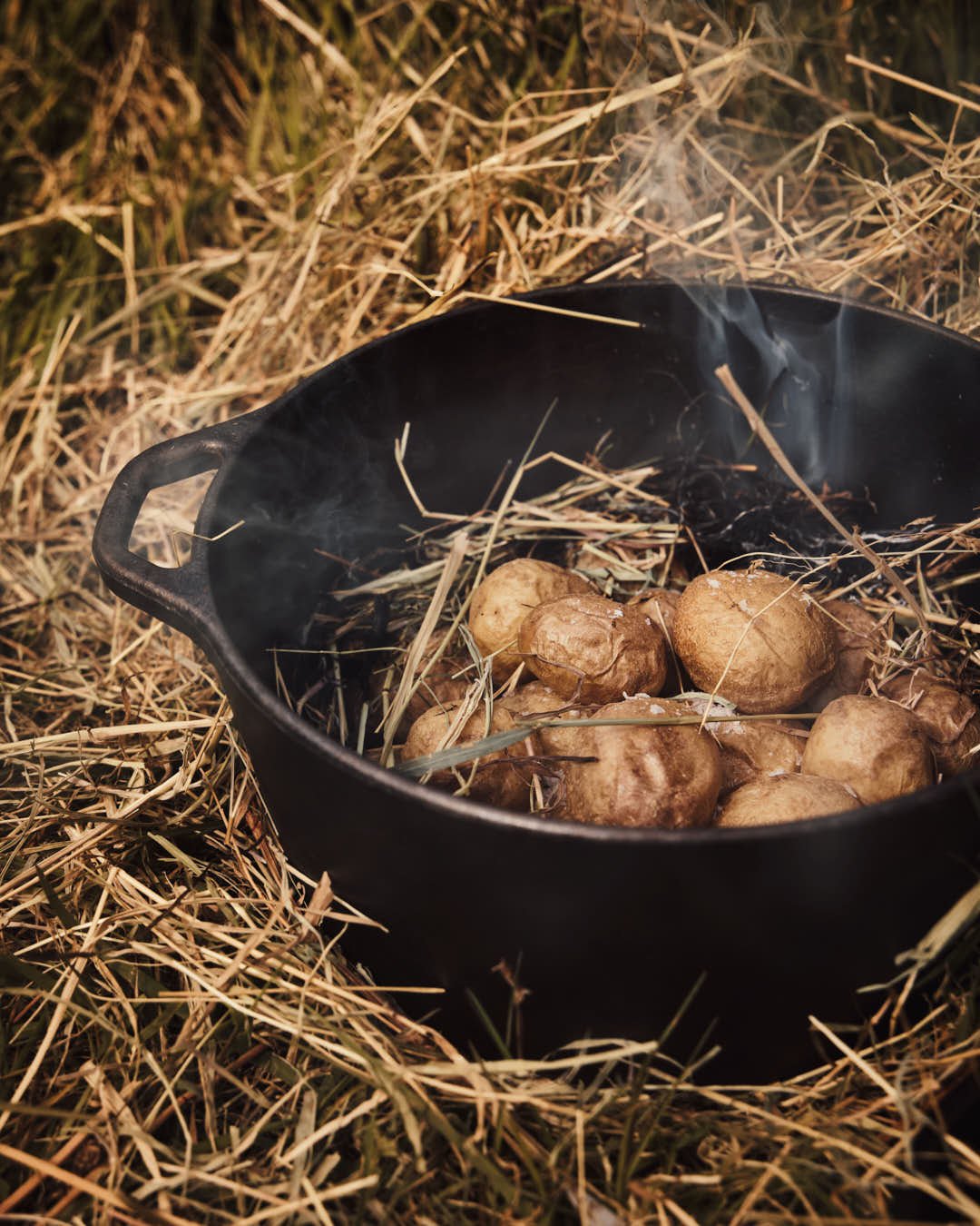 angled image of potatoes nestled in hay in cast iron pot with salt being sprinkled in outdoor setting