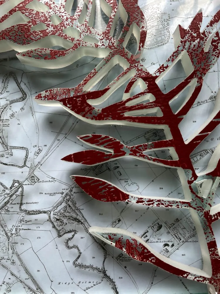 A layered image. The back plate glass has a decal or transfer image of the staiths in Gateshead. Lying on top of it is a waterjet cut out of a weed. The glass is 15mm in thickness and the top surface has an iron enamel fired on to it.