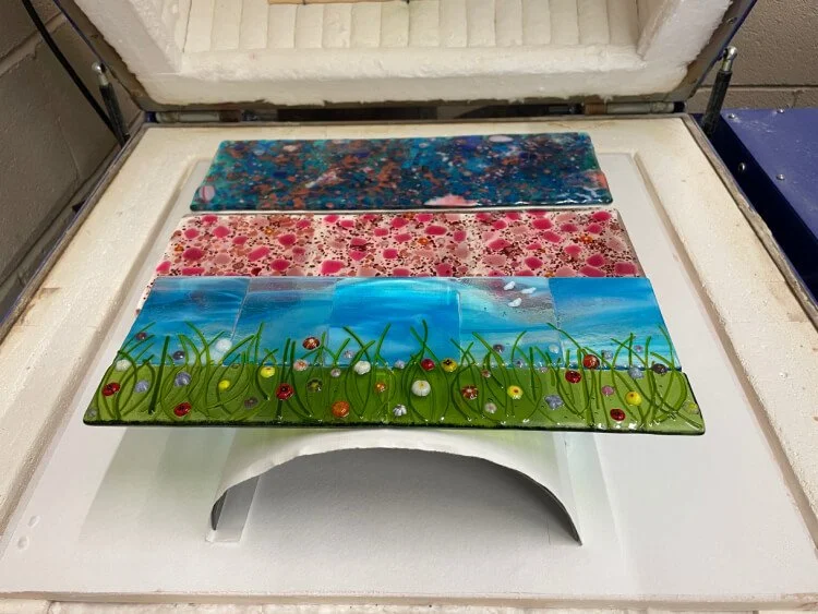 Two long pieces of glass are laid on top of two nibble dishes, ready for thme to be slumped. In front of them is a meadow scene laid on to of a metal curved mould.