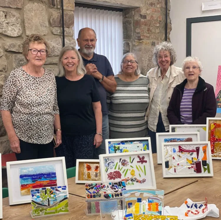 A group shot of the Creative age group with some of their finished work created over the past five weeks. Most of the pieces are in white frames. There are a few curved glass pieces in the front on the table. Glass Fusing. Workshop .