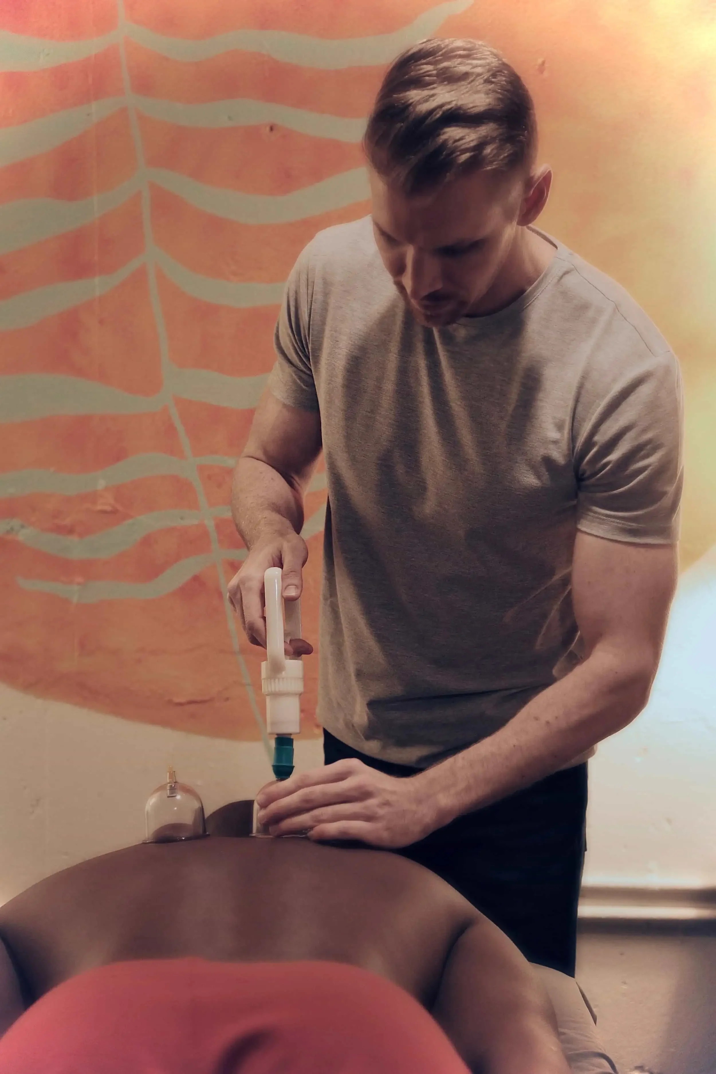 A man with short hair and a beige t-shirt performs acupuncture on a person lying face down. The background features a colorful abstract mural.