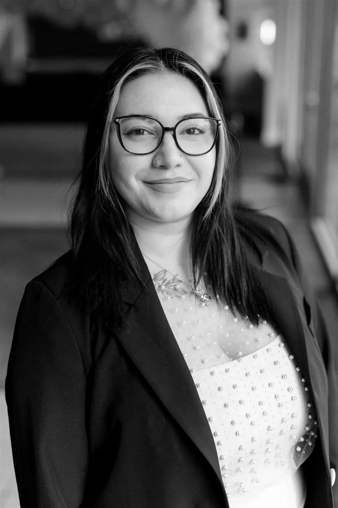 Black and white portrait of a woman with wavy hair, wearing glasses, smiling, and dressed in a black top with sheer, polka-dotted puffy sleeves, standing against a plain light background.