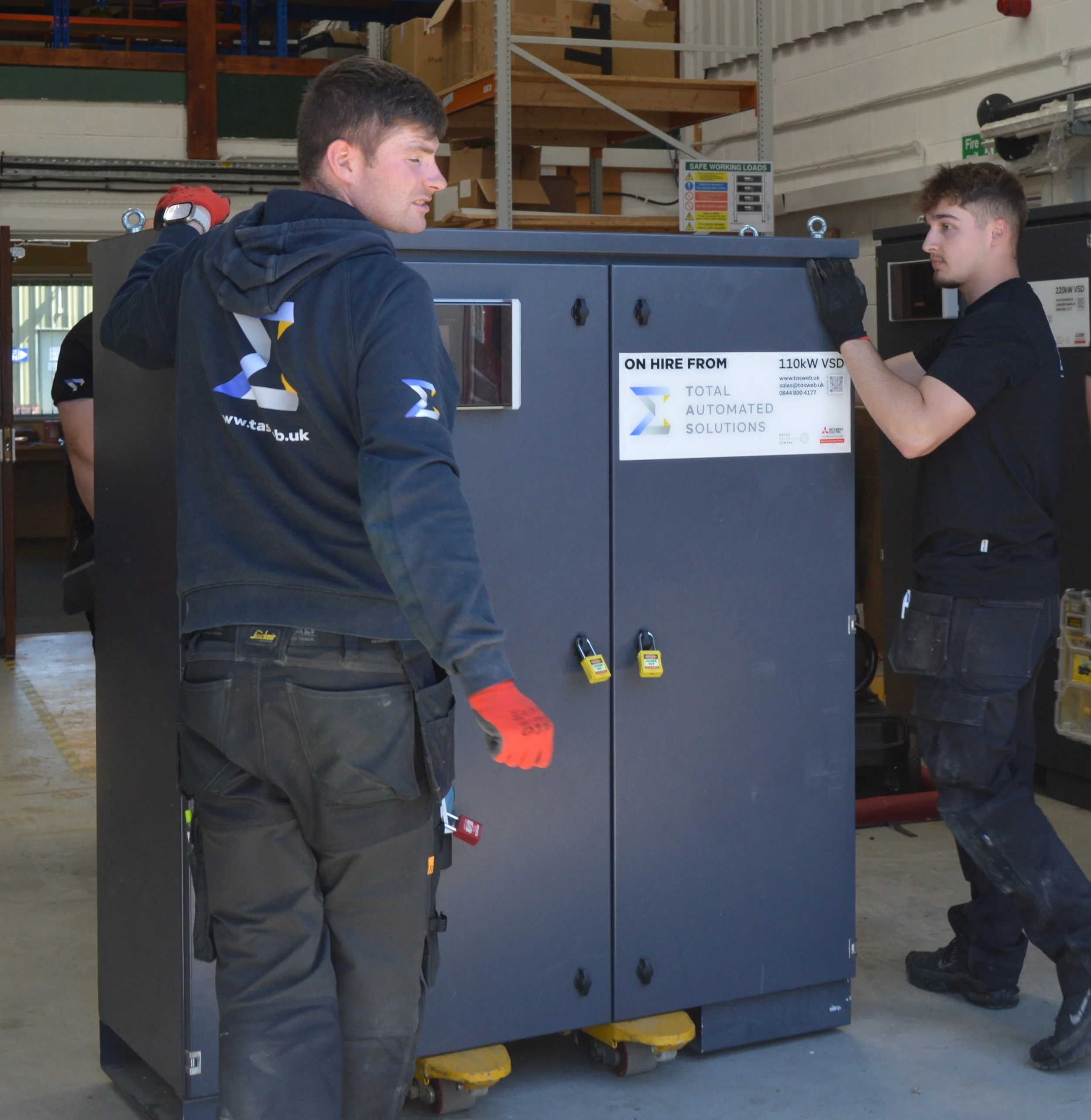 Two young men in a warehouse operating a large industrial machine with a control panel. Shelves with boxes and safety signs are visible in the background.