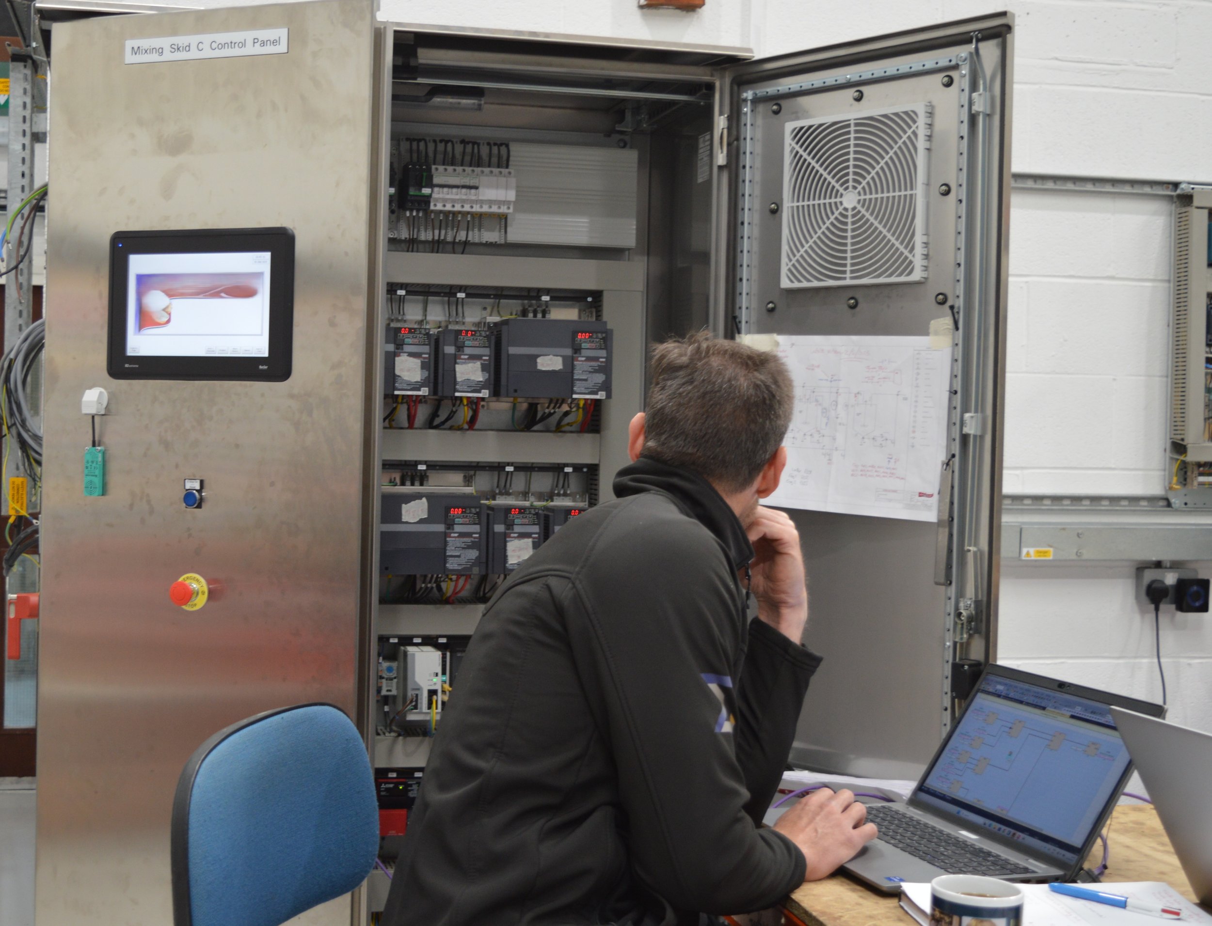 A person working with electrical control panels, using a laptop and observing a schematic diagram.