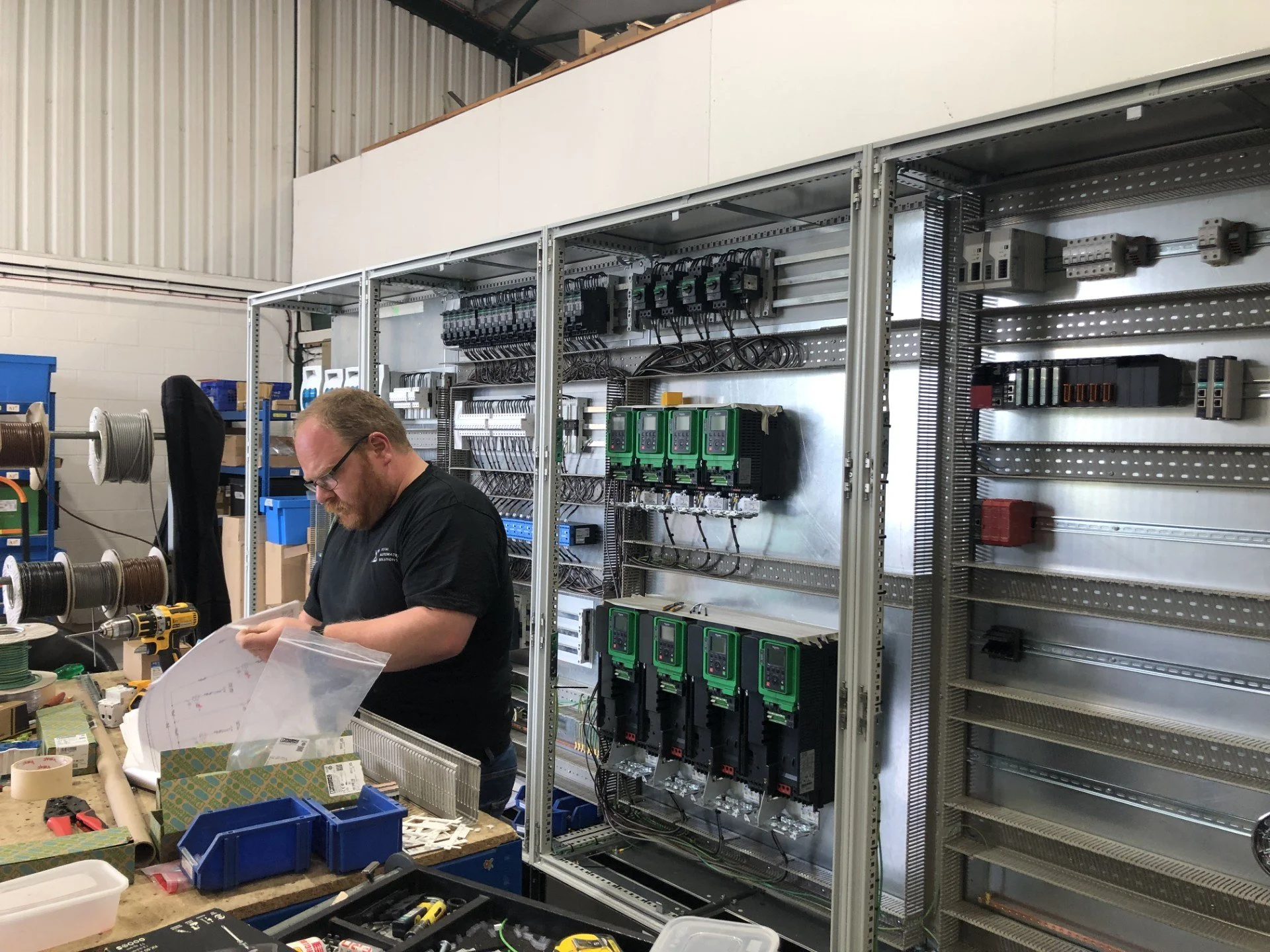 A man working on electronic components in a workshop with an open electrical control panel and shelves filled with various electronic devices and wires.
