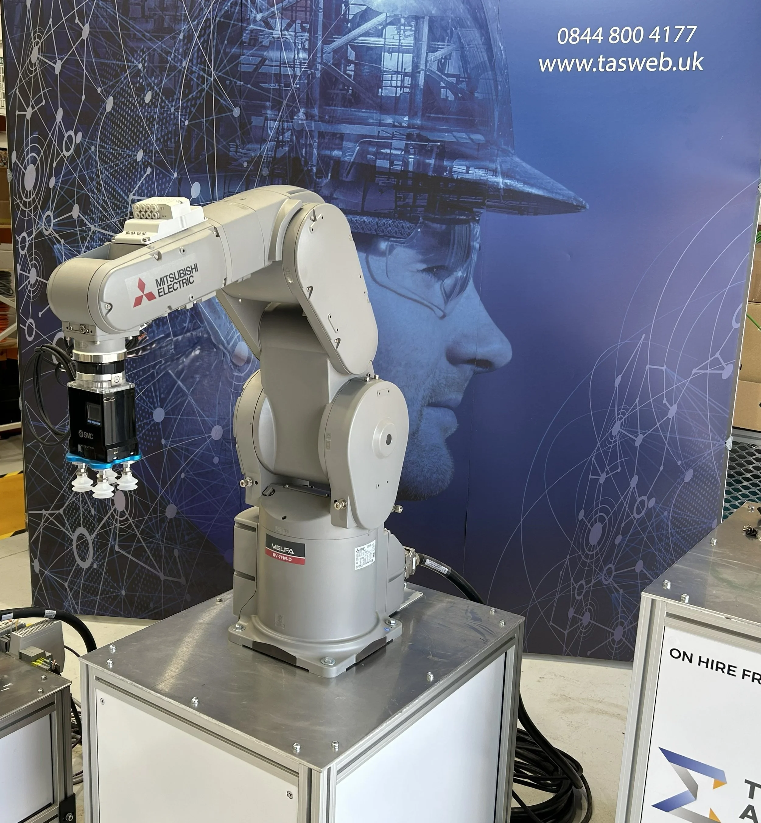 Industrial robotic arm labeled Mitsubishi Electric displayed on a metal table at a technology exhibition, with a backdrop featuring digital graphics and a human face outline.