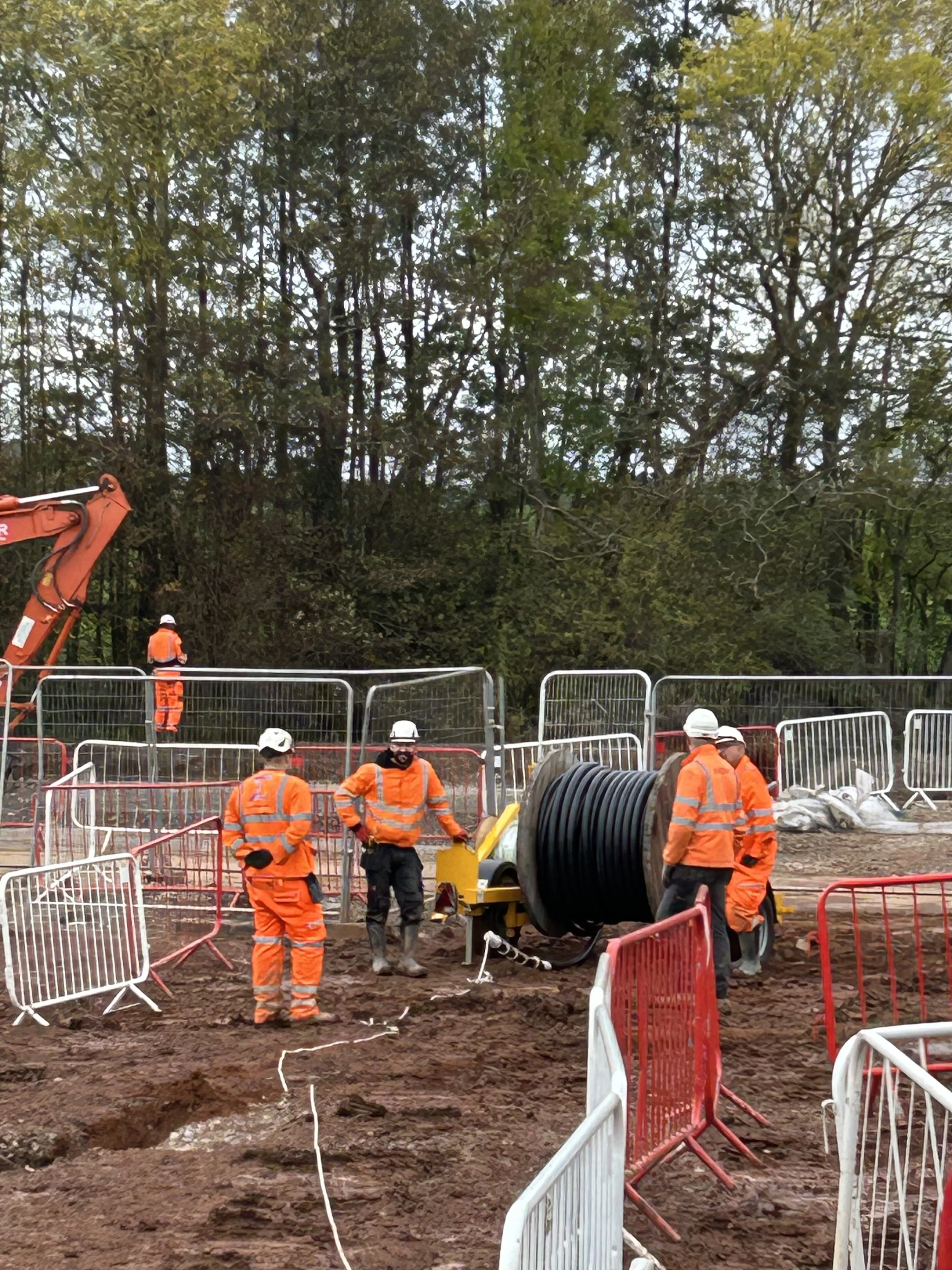 Construction workers in orange safety gear working on a site with fences, heavy machinery, and a large spool of cable, set outdoors with trees in the background.