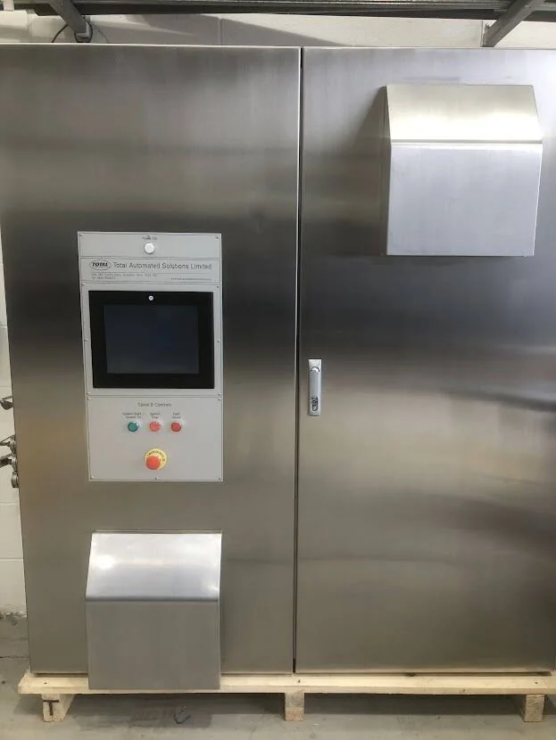 A stainless steel industrial control cabinet with a touchscreen, three buttons, and an emergency stop button, situated on a wooden pallet.