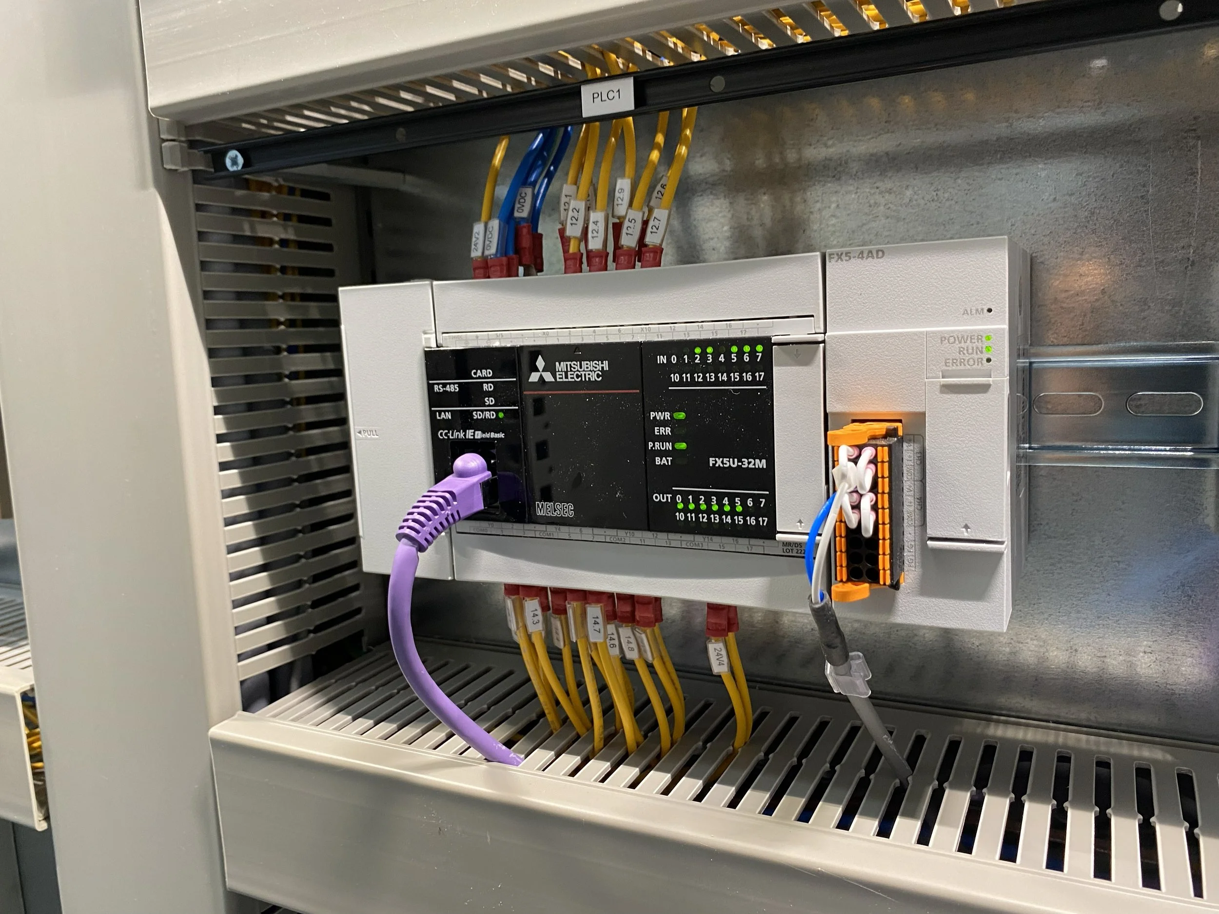 An industrial control panel with a Mitsubishi Electric programmable logic controller (PLC) and various colored wires connected to terminal blocks.