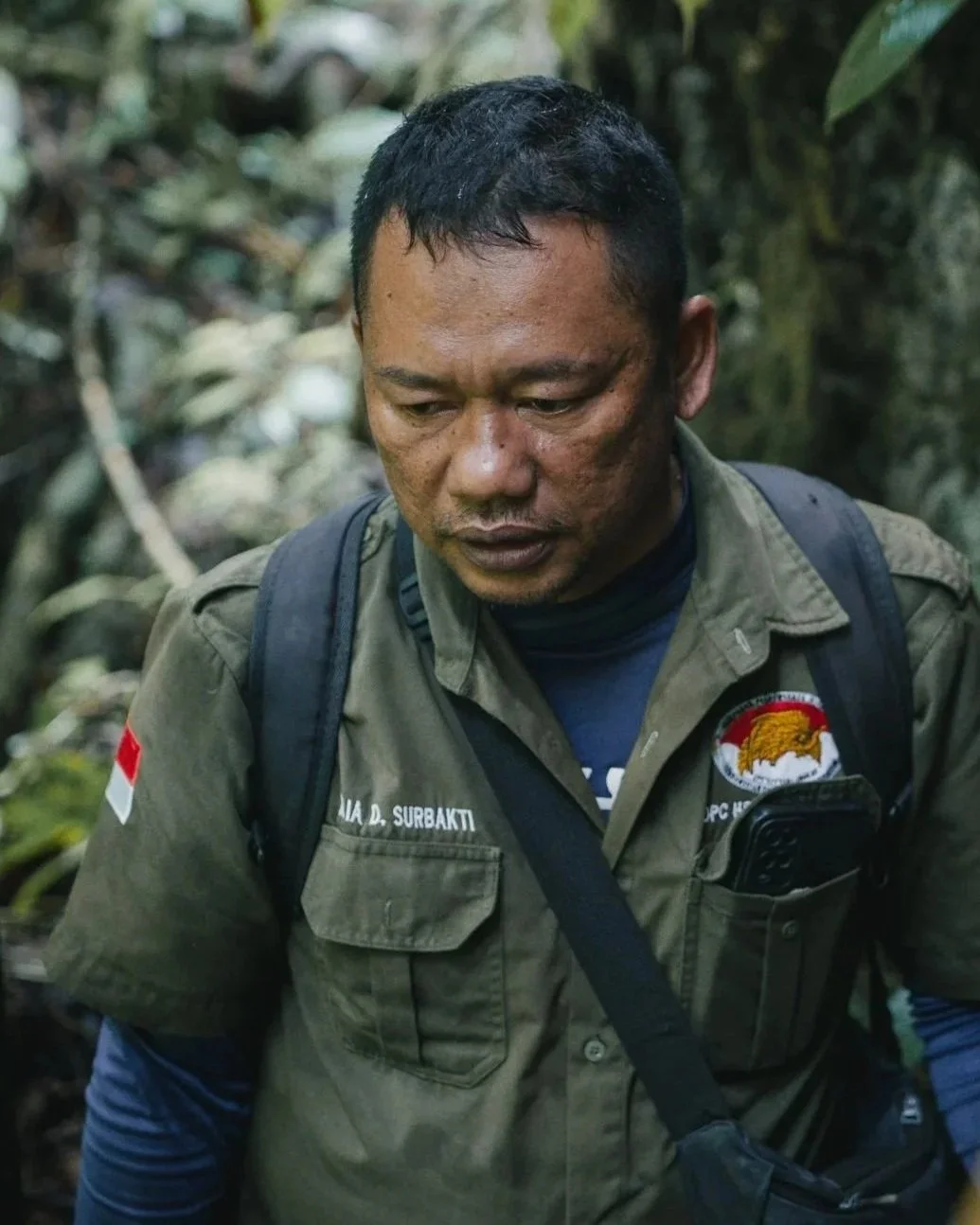 A man wearing a green outdoor shirt with patches, a backpack, and a waist strap, looking downward in a dense forest.