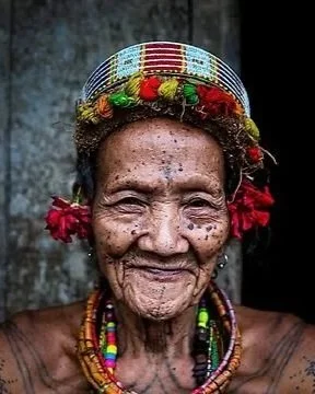 An elderly woman with traditional jewelry, including a beaded necklace and a decorative headband, smiling against a dark background.