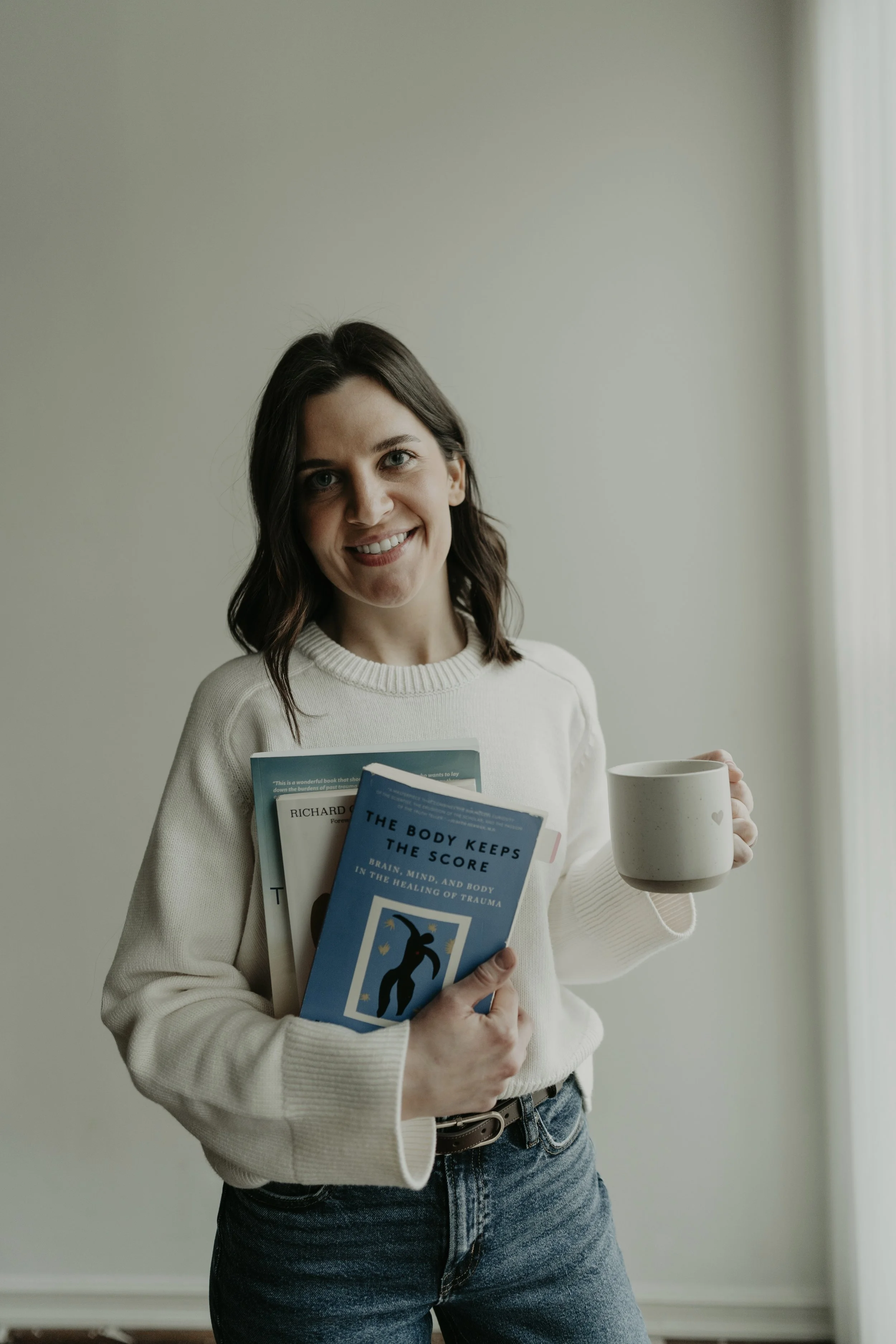 Therapist holding books about trauma, Internal Family Systems (IFS), and emotional healing while holding a coffee mug.