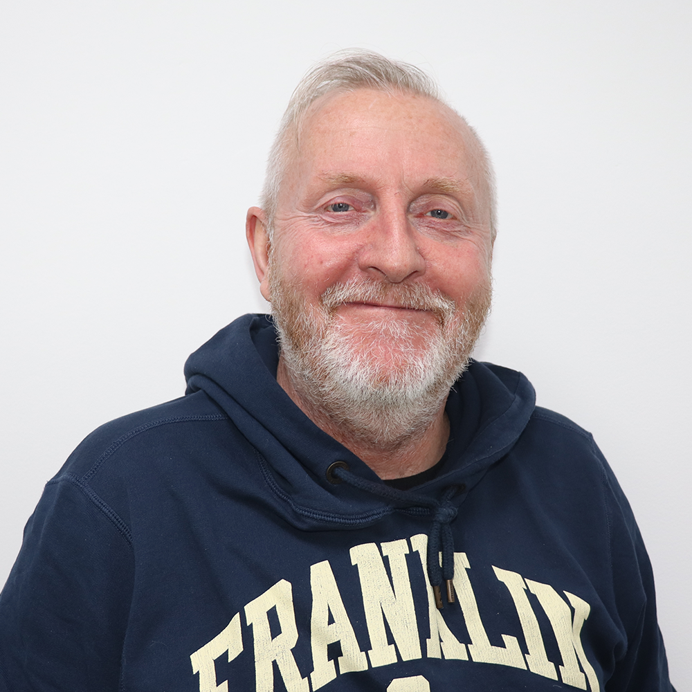 A smiling older man with white hair and a beard, wearing a navy hoodie with the word 'FRANKLIN' written on it, standing against a white background.