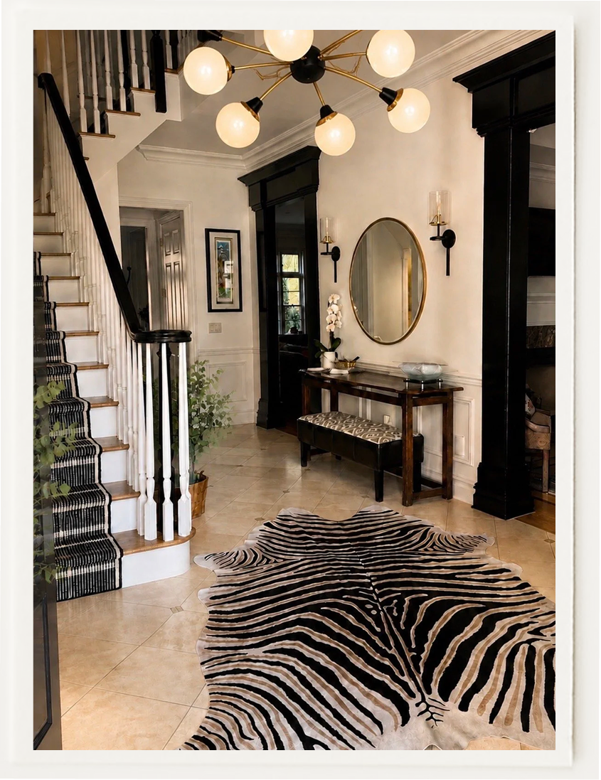 Elegant entryway with a black-and-white staircase, zebra-print rug, oval mirror, console table, and globe chandelier