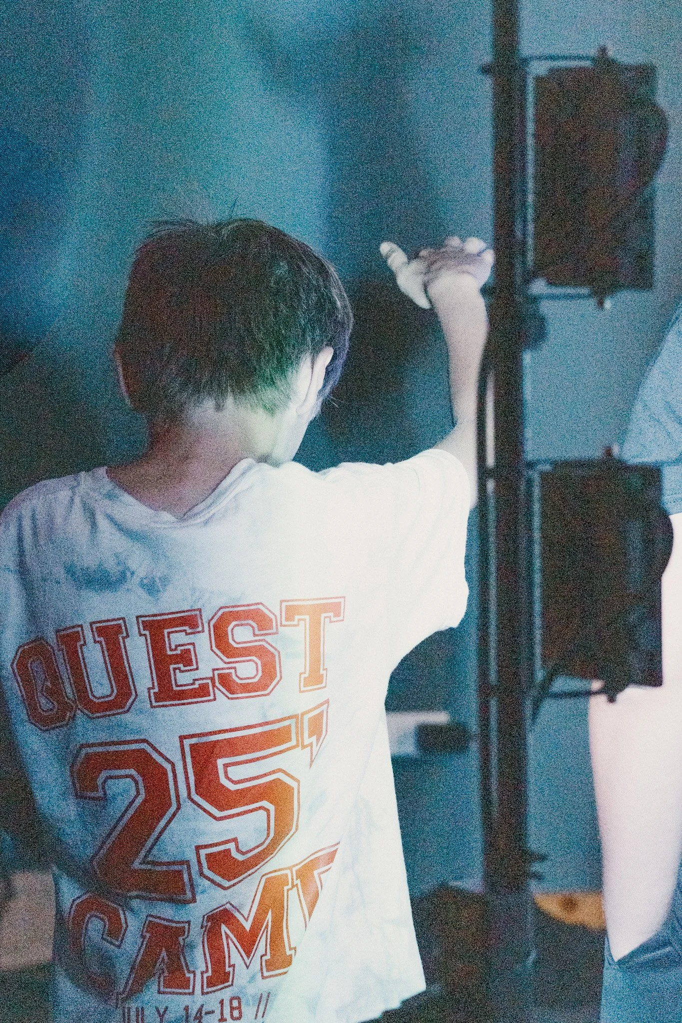 A person with short dark hair seen from behind, wearing a white T-shirt with red and orange lettering, reaching out with their right hand, standing near black speaker equipment in a dimly lit setting.