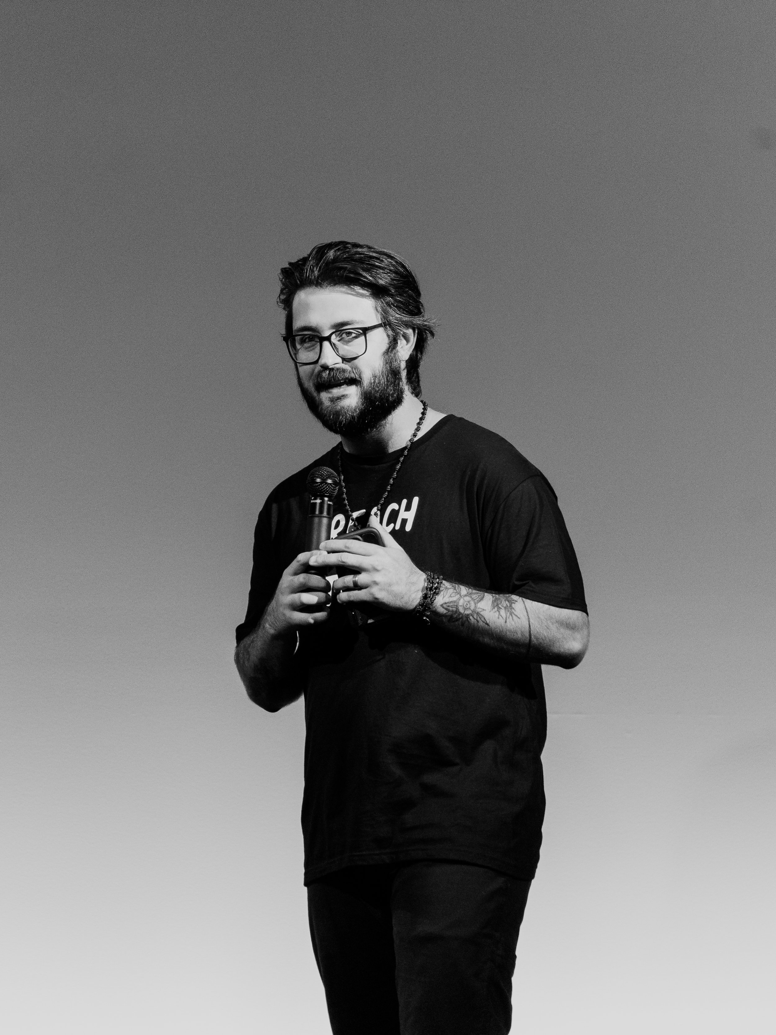 A man with dark hair, glasses, and a beard holding a microphone and speaking on stage, wearing a black t-shirt with white text, against a plain background.