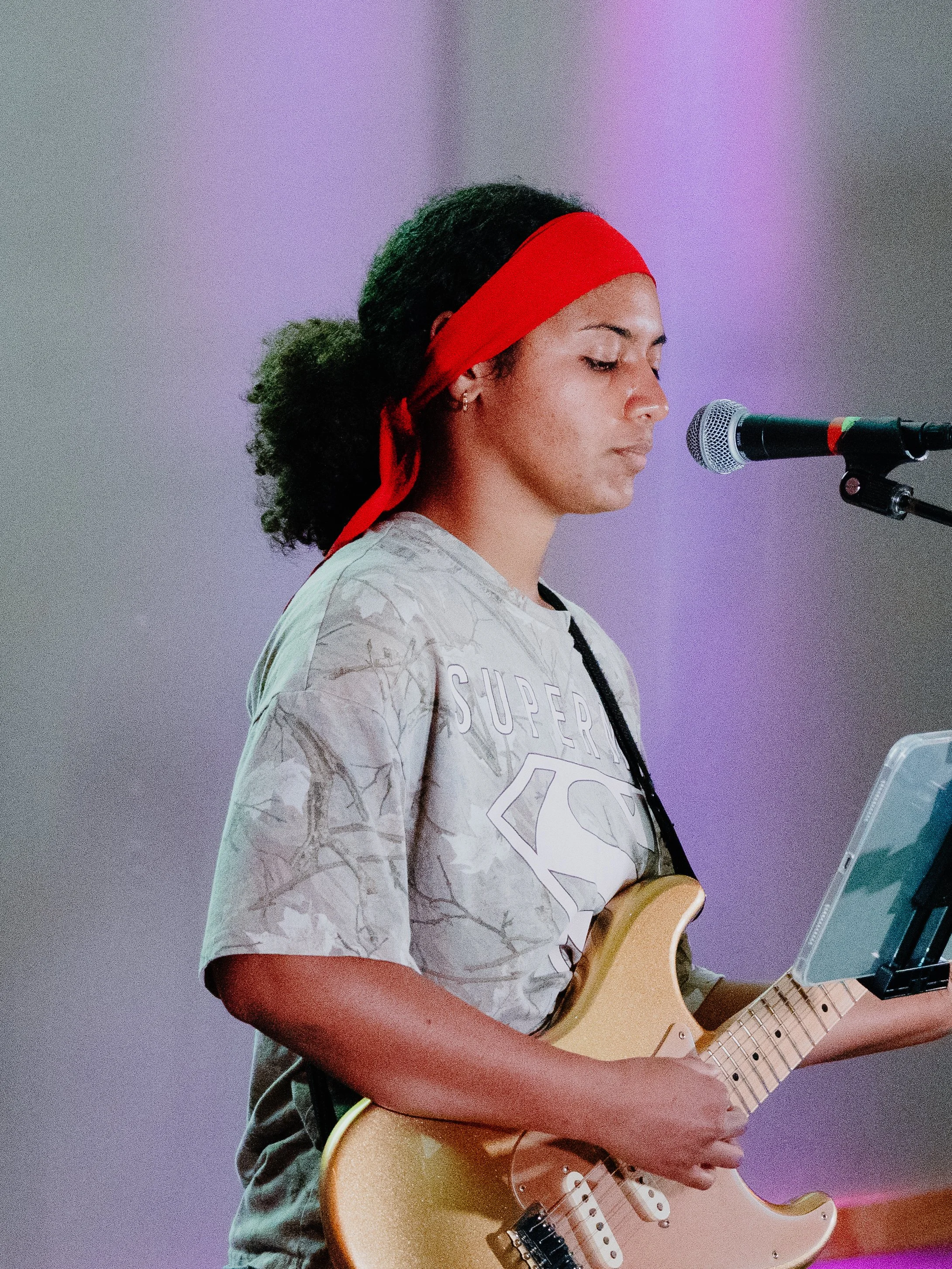 A young woman with curly hair tied back with a red headband playing an electric guitar and singing into a microphone.