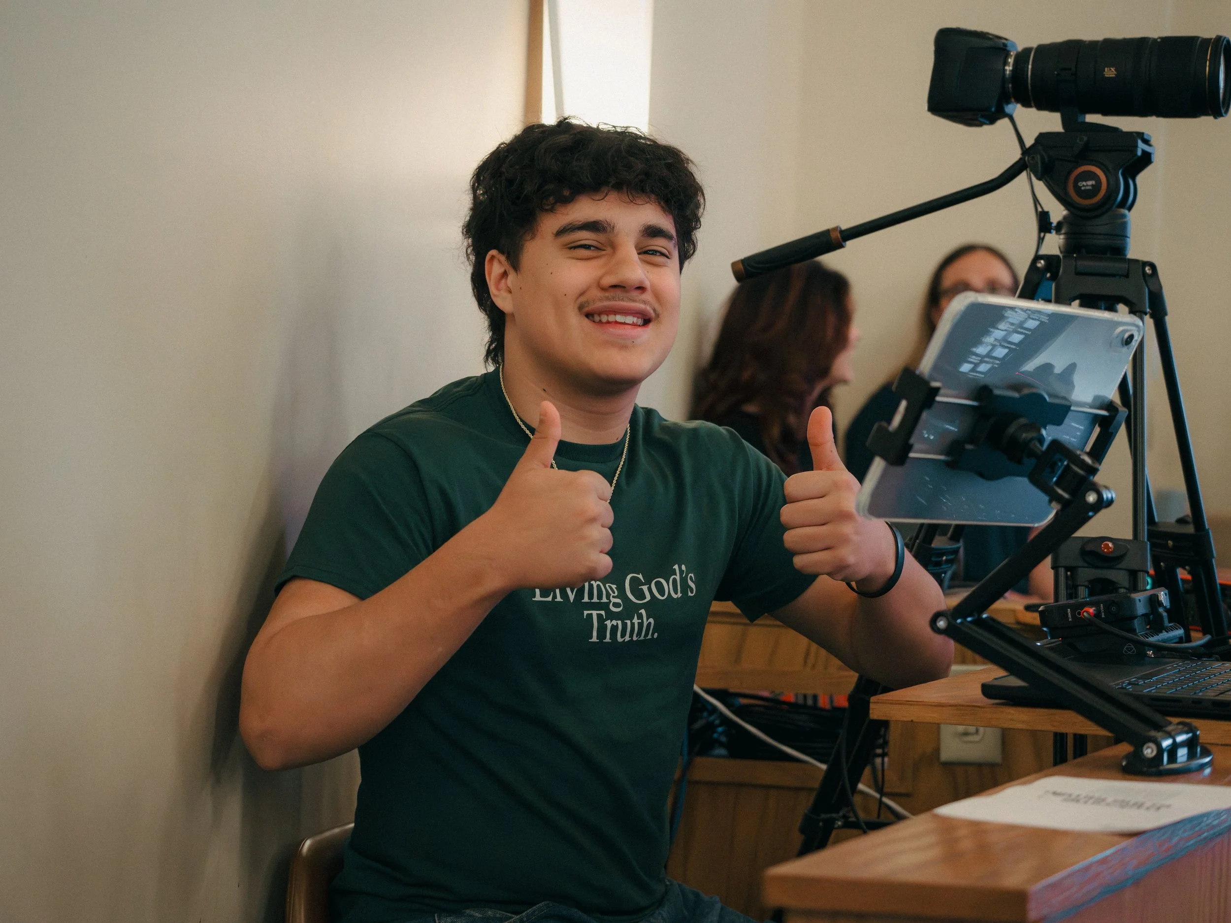 A young man with curly dark hair smiling and giving thumbs up while sitting at a table with camera equipment, including a large camera mounted on a tripod, in a room with neutral-colored walls.