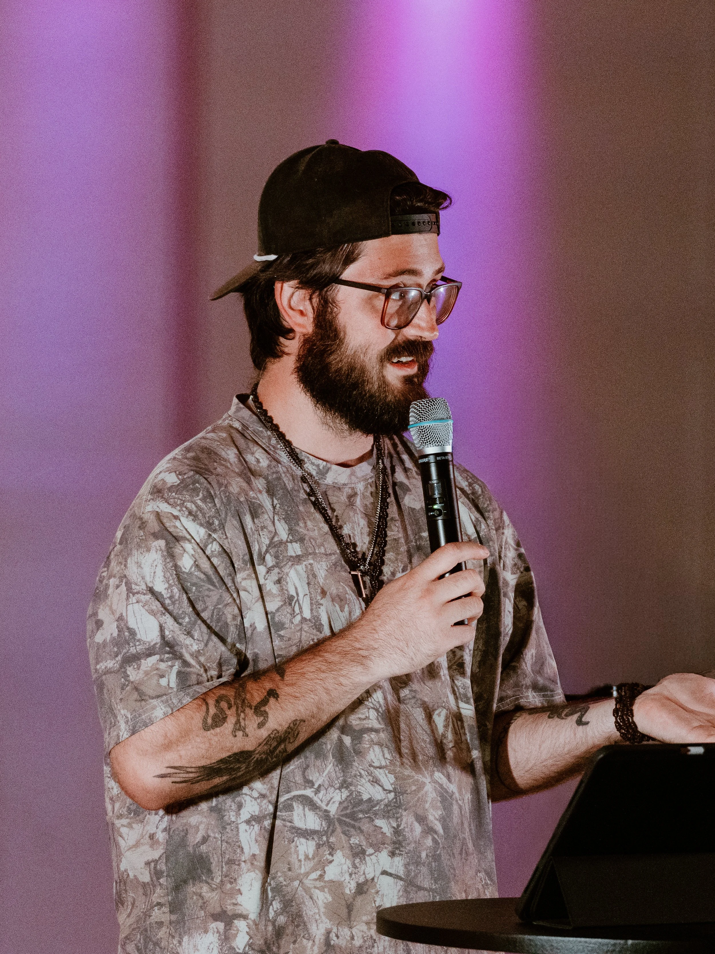 A man with glasses, a beard, and tattoos on his arms, wearing a black cap and a patterned T-shirt, speaking into a microphone in front of a purple background.