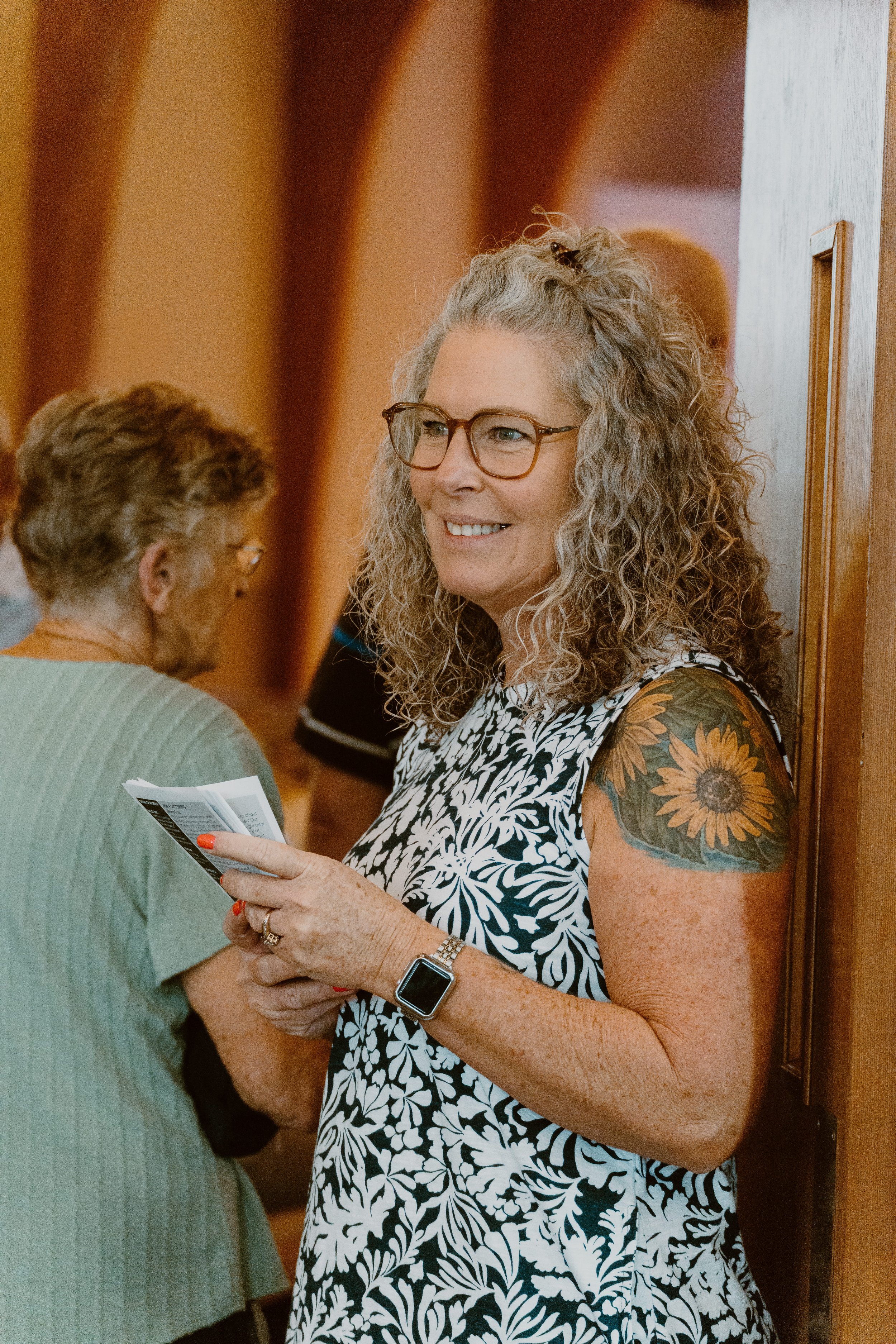 A woman with curly gray hair, glasses, and a sunflower tattoo on her upper arm, smiling and holding a pamphlet while talking to an elderly woman in a gray sweater in a hallway.