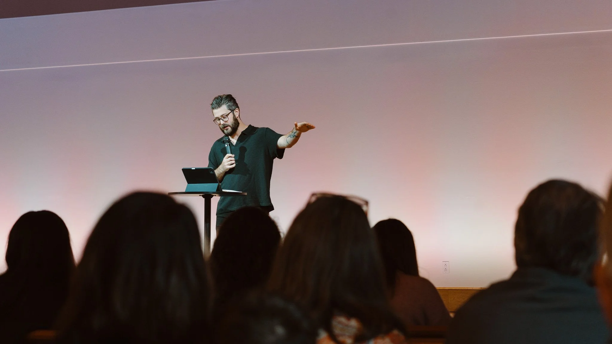 A man standing at a small table on stage giving a presentation, holding a microphone in one hand and gesturing with the other, with an audience sitting in front of him.