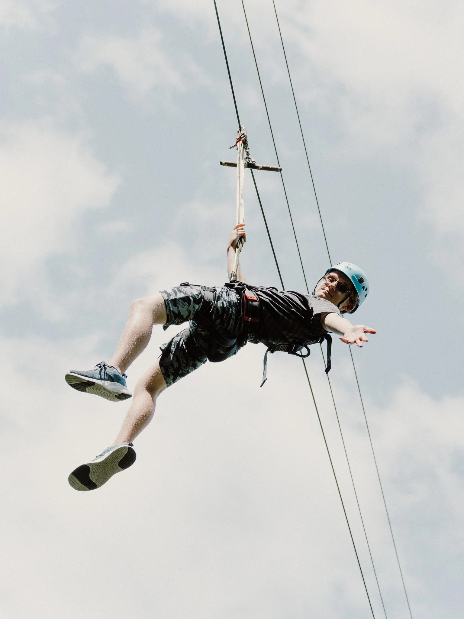A person wearing a safety helmet and sunglasses is riding a zip line against a cloudy sky, holding onto the zip line with one hand and extending the other hand outward.