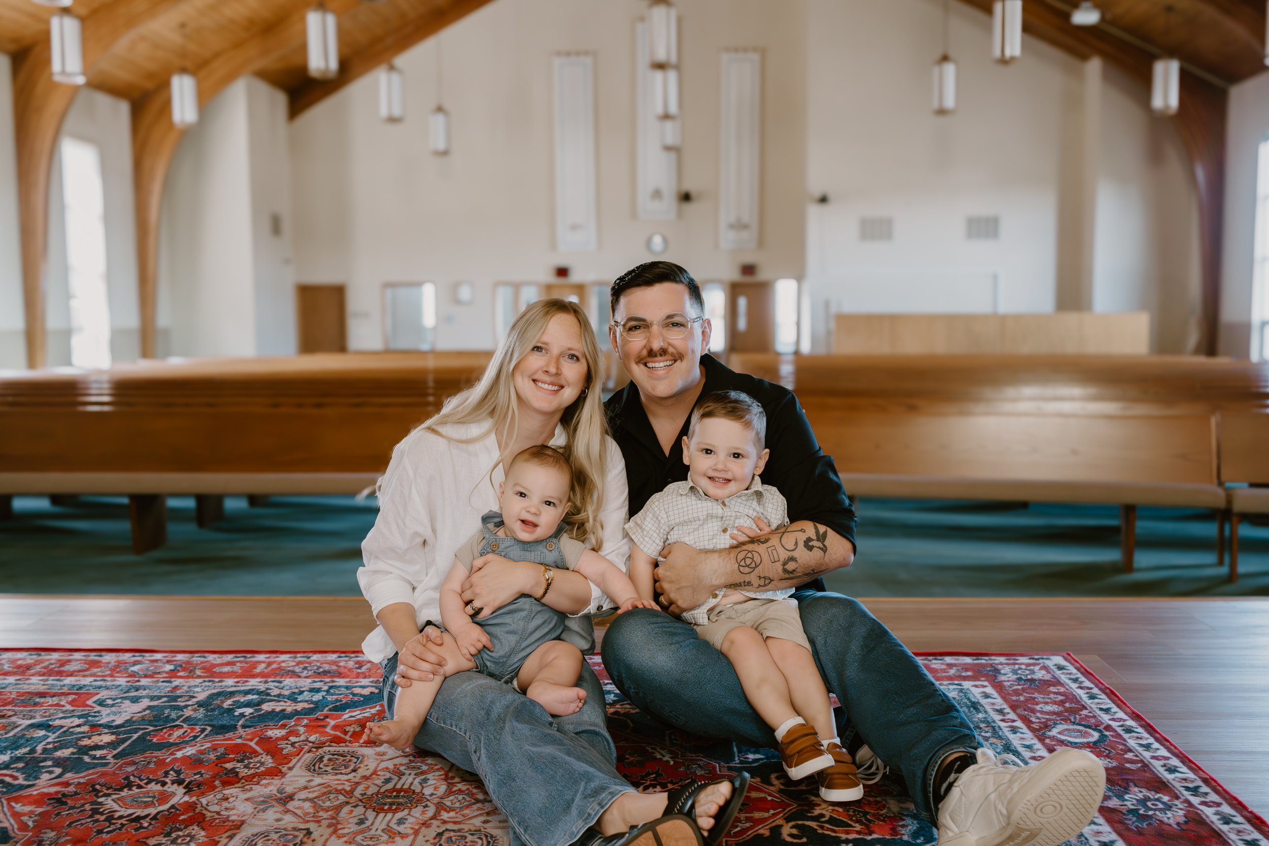 A family of four sitting on a patterned red and blue rug inside a church. The mother has long blonde hair and is wearing a white shirt. The father has short dark hair, glasses, a mustache, tattoos on his right arm, and is wearing a black shirt. The daughter has light hair and is wearing a denim dress. The son has short hair, a checkered shirt, and khaki shorts. They are smiling and looking at the camera.