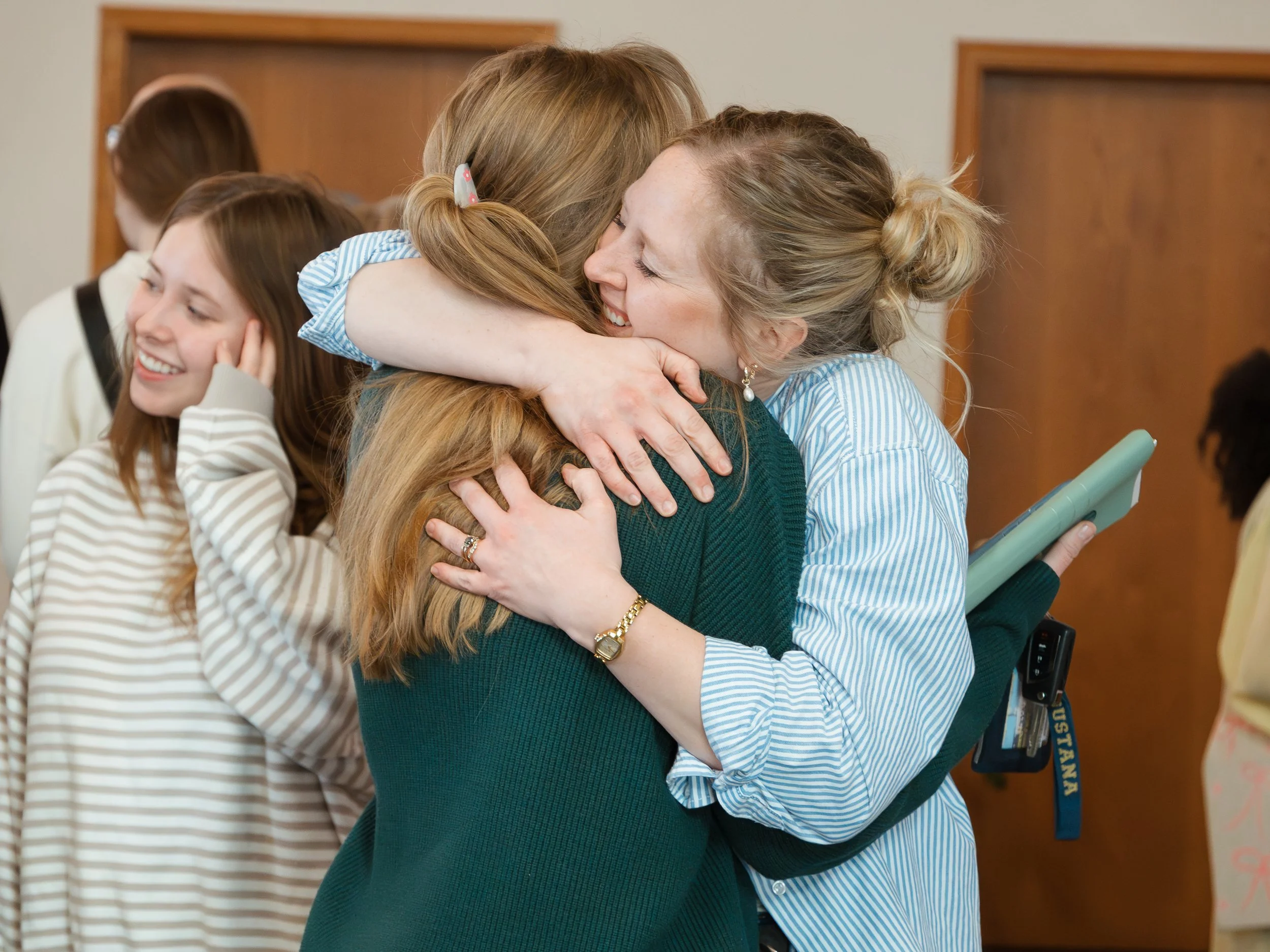 Two women hugging and smiling at each other in a room with other people in the background.