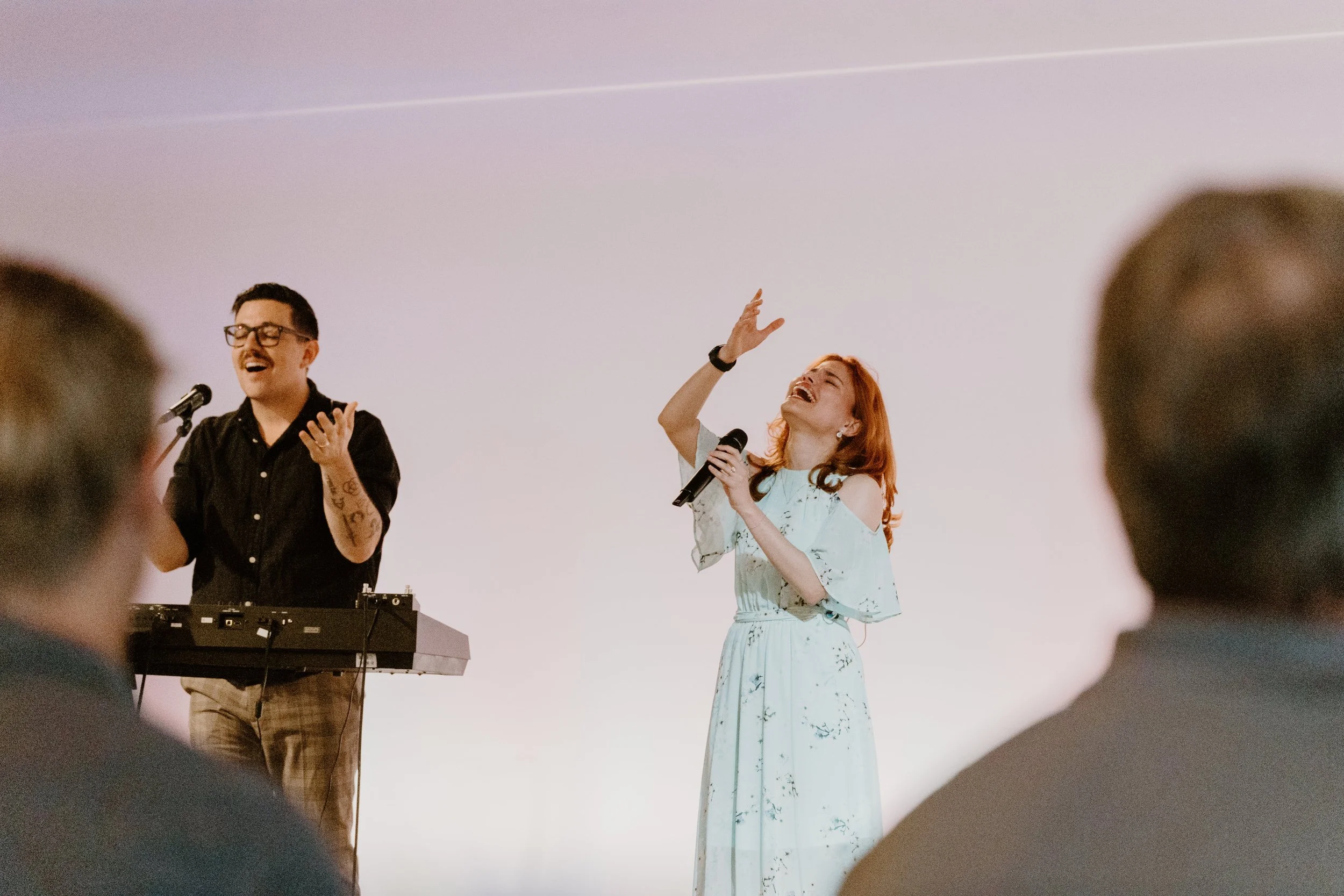 A man and woman singing and performing on stage in front of an audience, with the woman holding a microphone and both looking joyful.