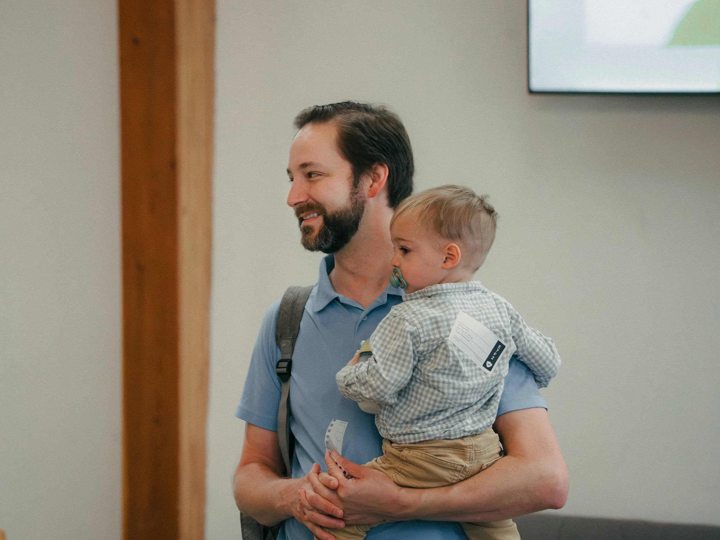 A man with dark hair and a beard holding a young child wearing a plaid shirt and beige pants. They are indoors, with a wall and a screen in the background.