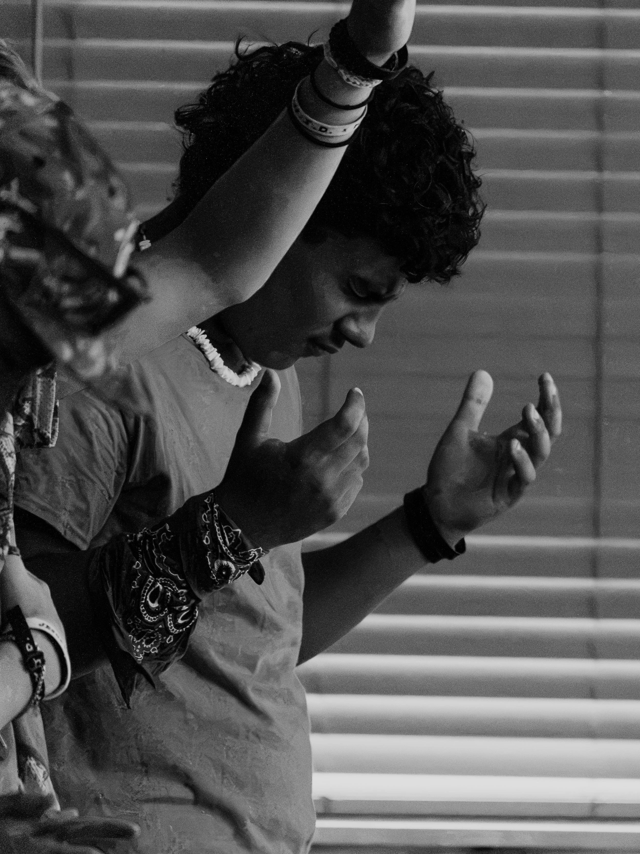 A person with curly hair, wearing jewelry and casual clothing, is seen with their hands raised and fingers pointing, in front of horizontal blinds.