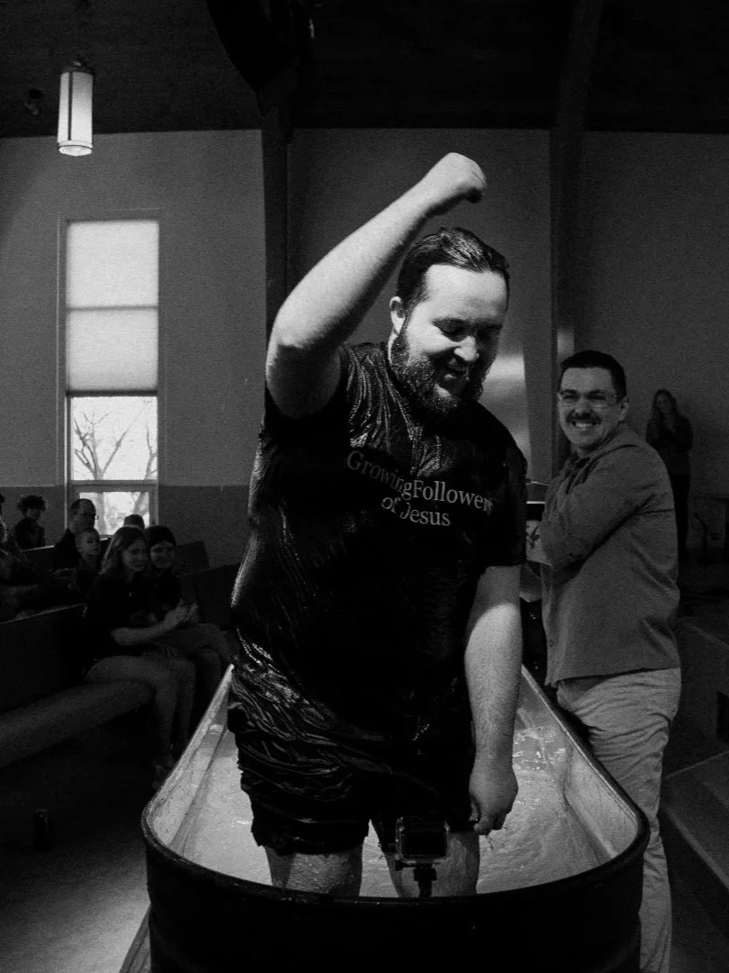 Man standing in a baptism pool with water, raising his right arm and smiling, with guests seated in the background inside a church.