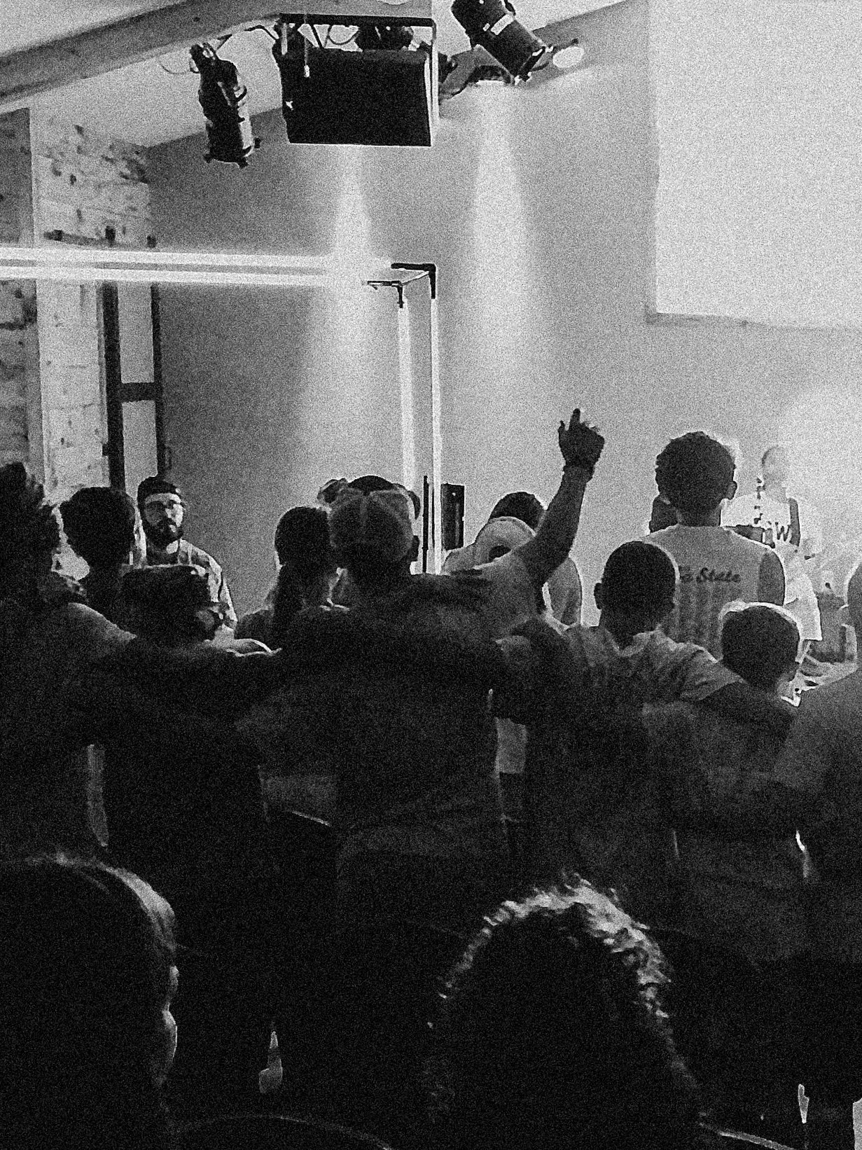 A black-and-white photo of a crowd attending a presentation or lecture in an indoor space with stage lighting, a screen, and a man raising his hand.