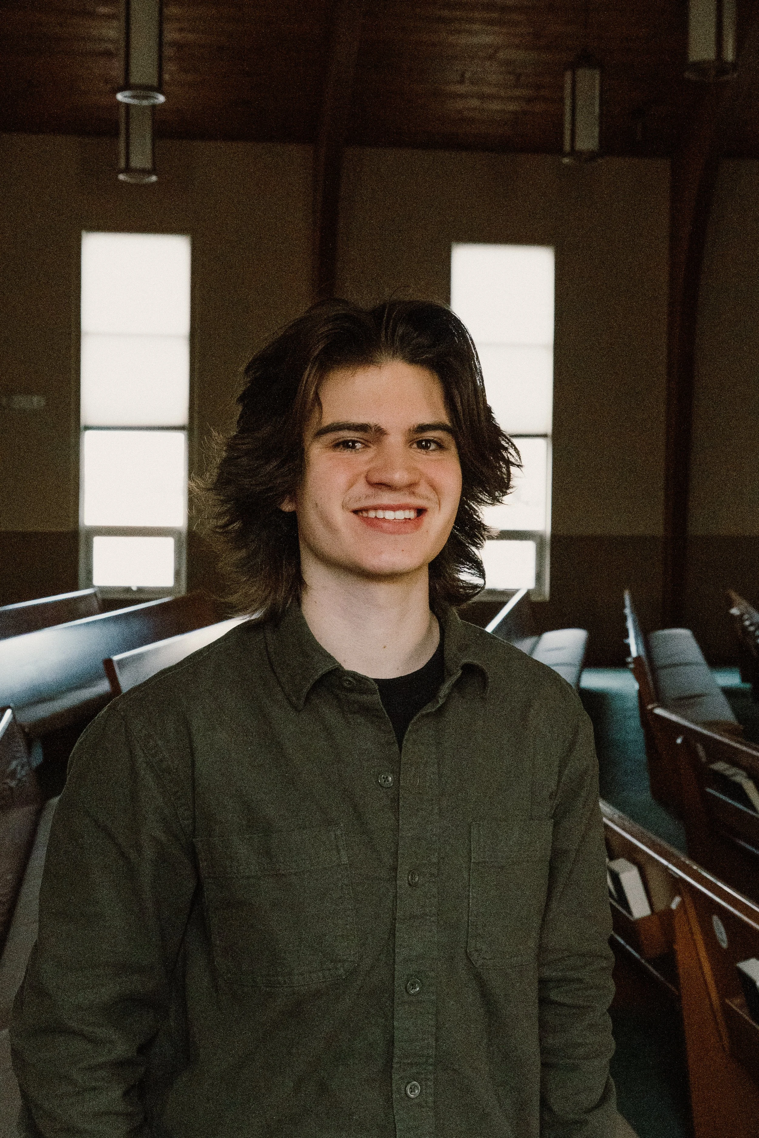 Young man with dark, wavy hair smiling, wearing a dark green button-up shirt, standing in a room with wooden ceiling beams and church pews.