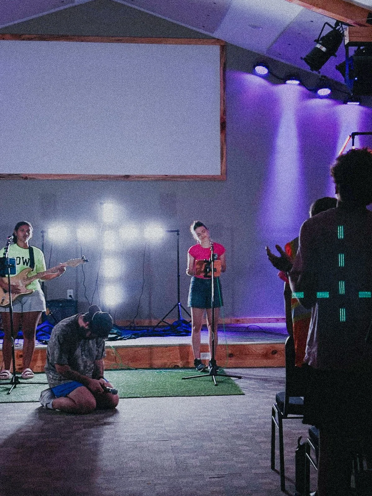 People performing on stage with musical instruments and microphones, purple stage lighting, and an audience member clapping.