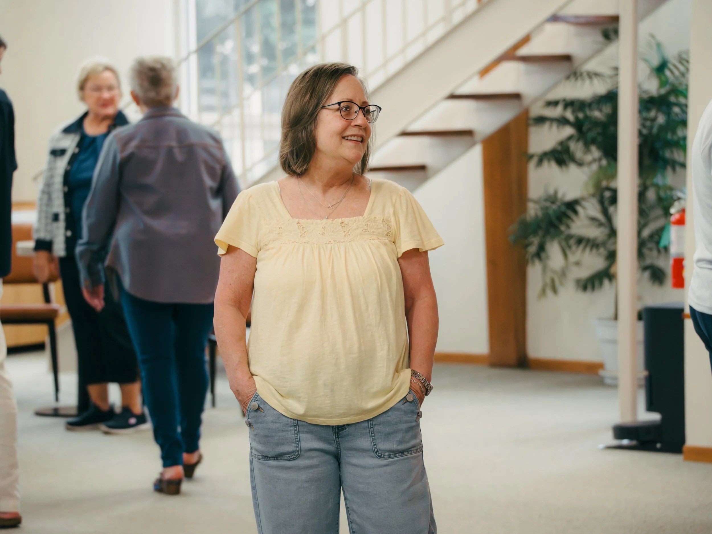 Smiling woman wearing glasses and a yellow blouse standing in a hallway with people in the background.