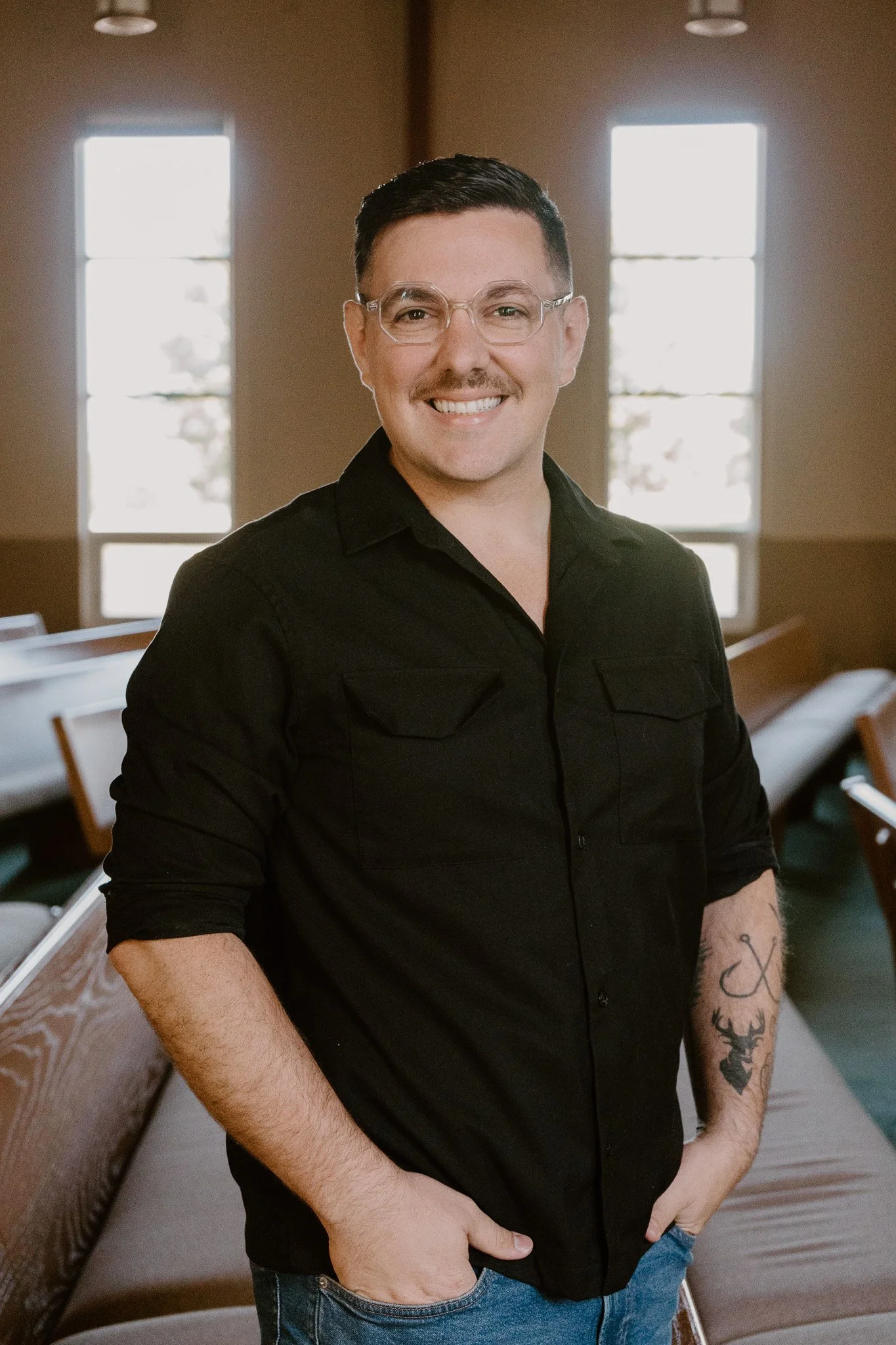 A smiling man with glasses and a mustache standing inside a church or chapel, with large windows in the background. The Reverend Zachary Fraley, MDiv. Lead Pastor at Christ's Family Church.