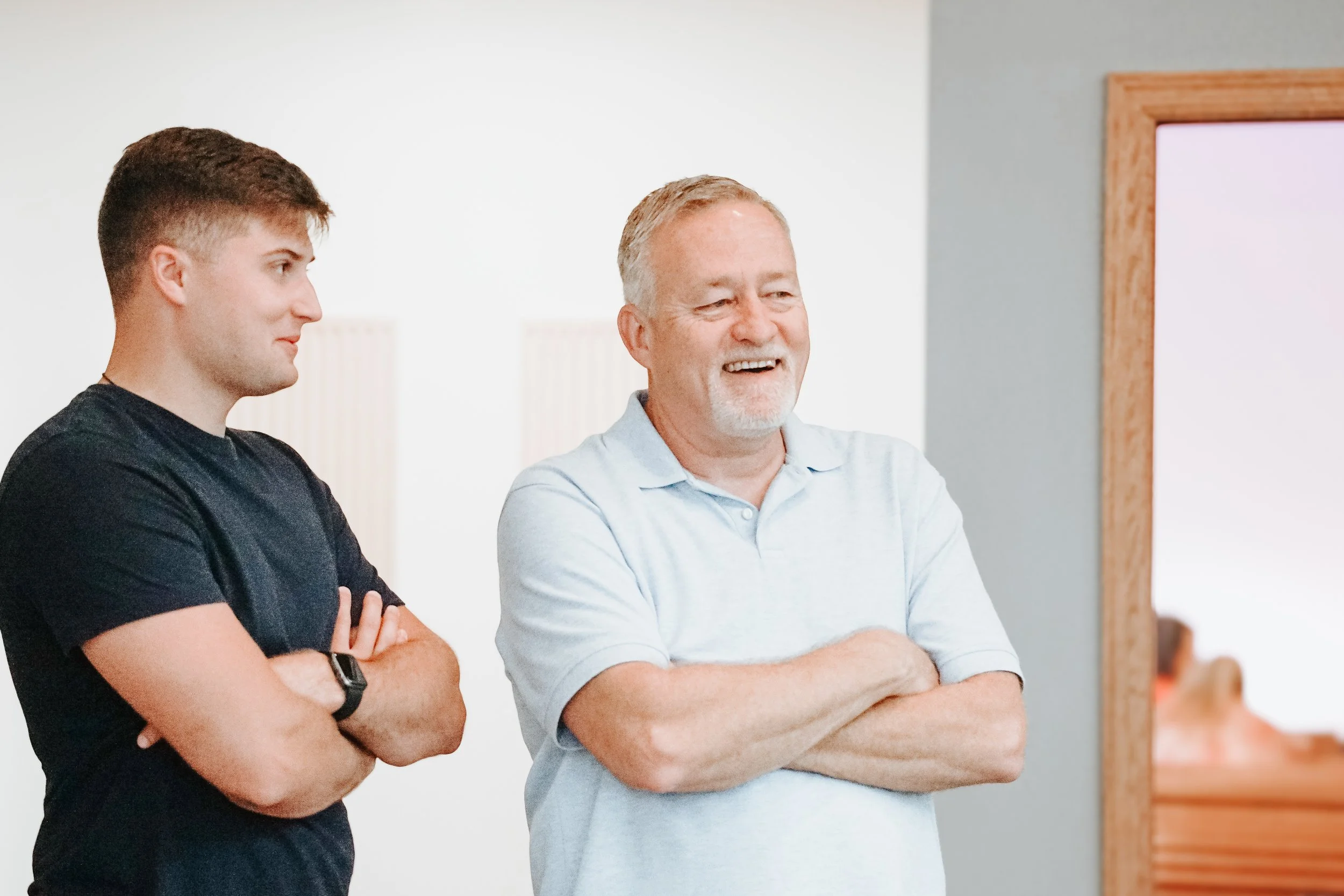 Two men standing with arms crossed, smiling and looking to the side, in an indoor setting.