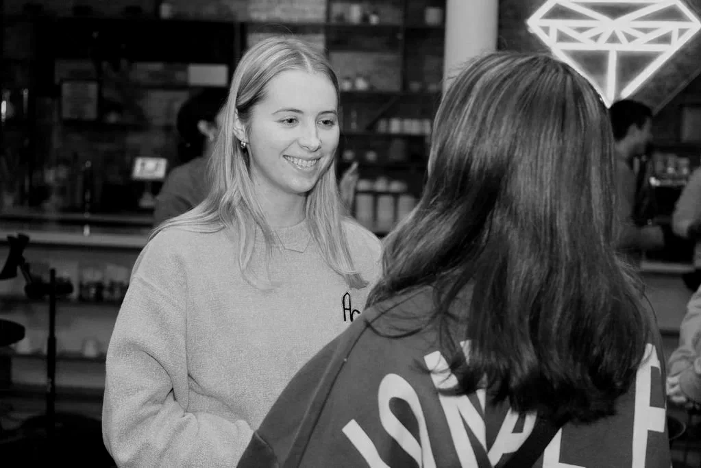 Two women talking in a cozy cafe or restaurant, with shelves and neon decor in the background.