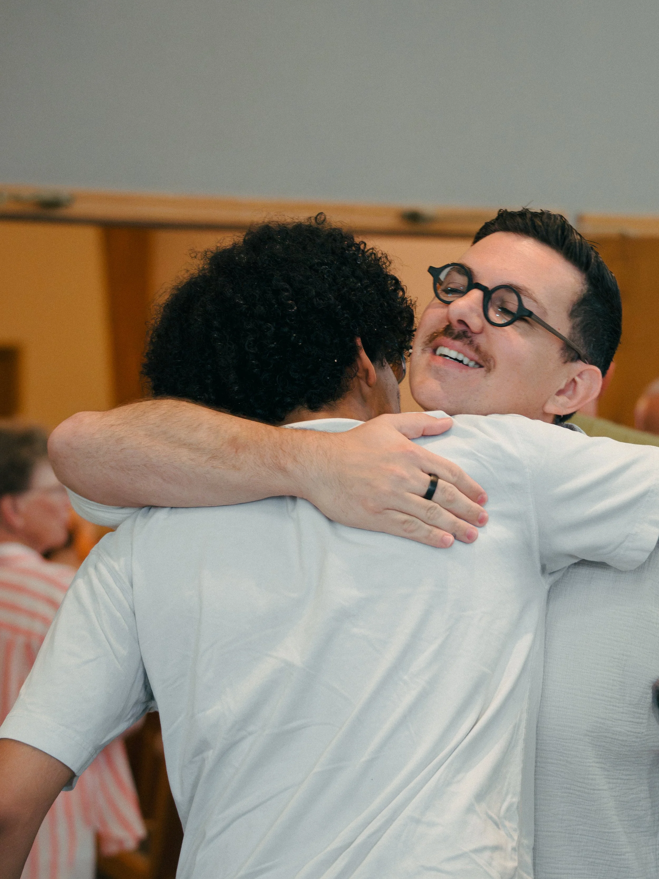Two men hugging warmly, smiling, and embracing in a close-up shot at a social gathering.