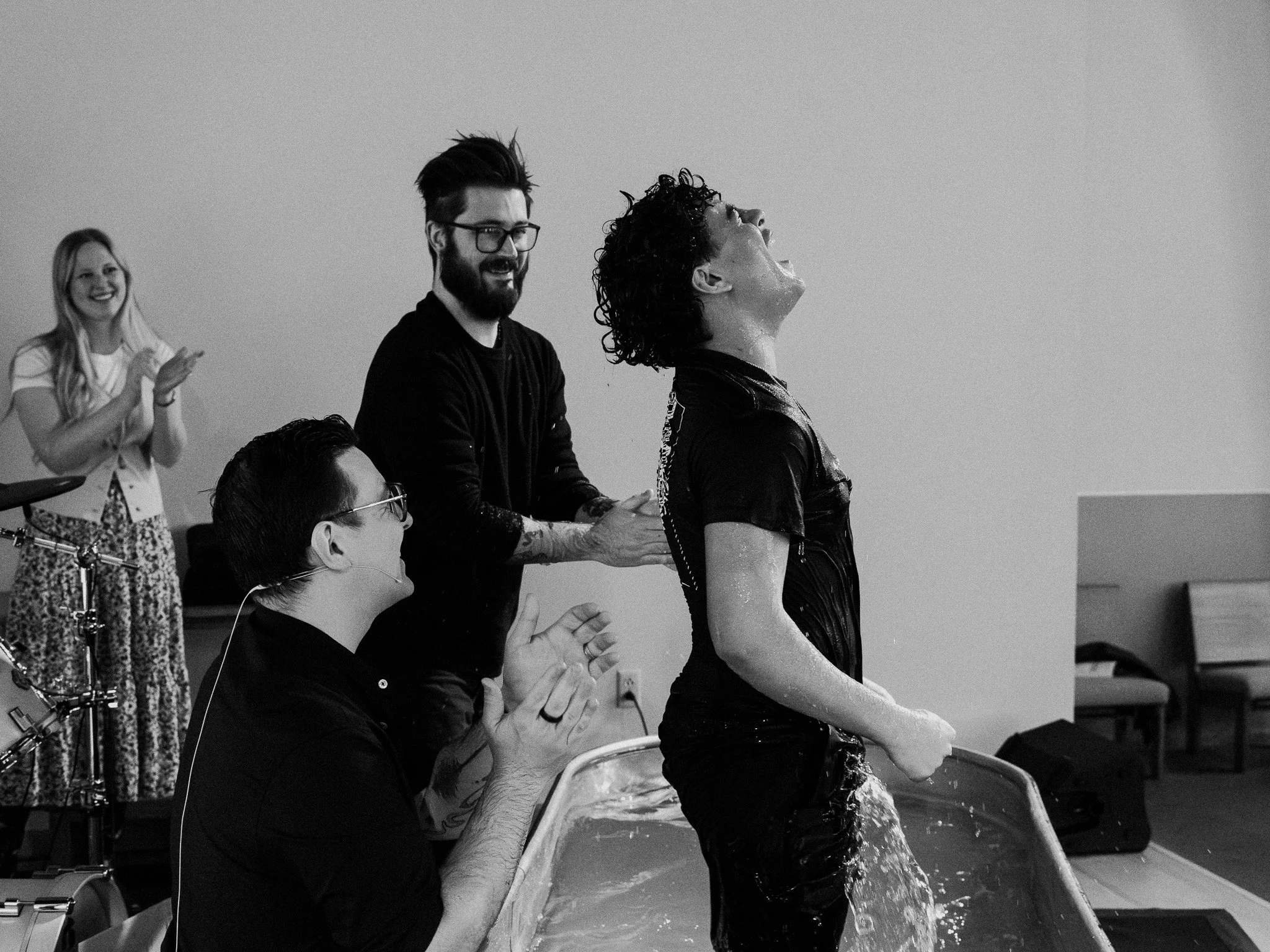 A group of people, some clapping and smiling, watching a woman with short curly hair pour water over her head at what appears to be a baptism or water ceremony. The woman is wet and dressed in black, and others are seated, observing and clapping in a room with a plain wall.
