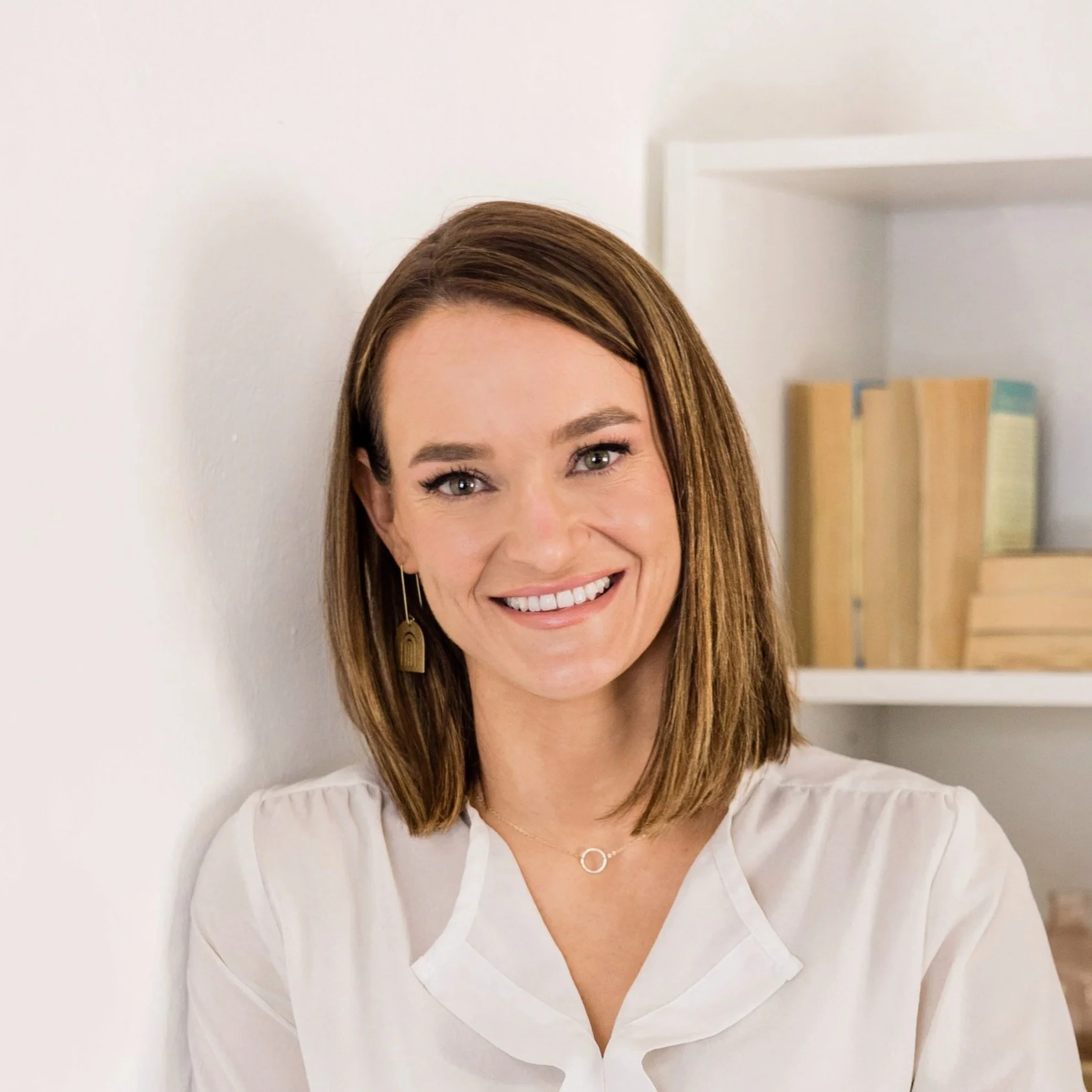 A woman with shoulder-length brown hair, smiling, wearing gold earrings and a white blouse, in a room with bookshelves in the background.