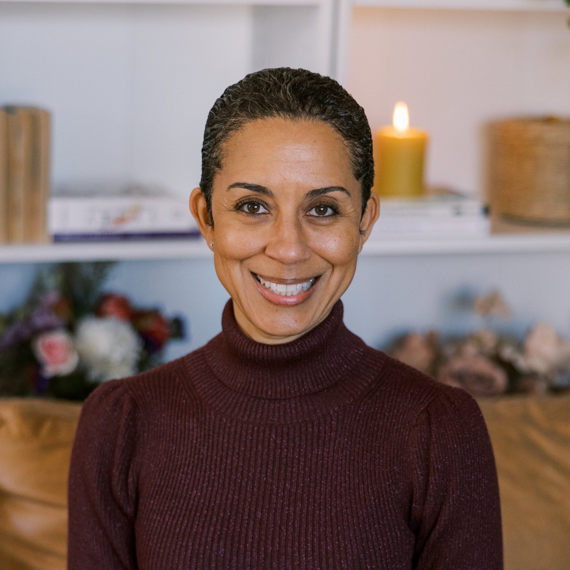 A smiling woman with short dark hair wearing a maroon turtleneck sweater indoors, with a candle, books, and flowers in the background.